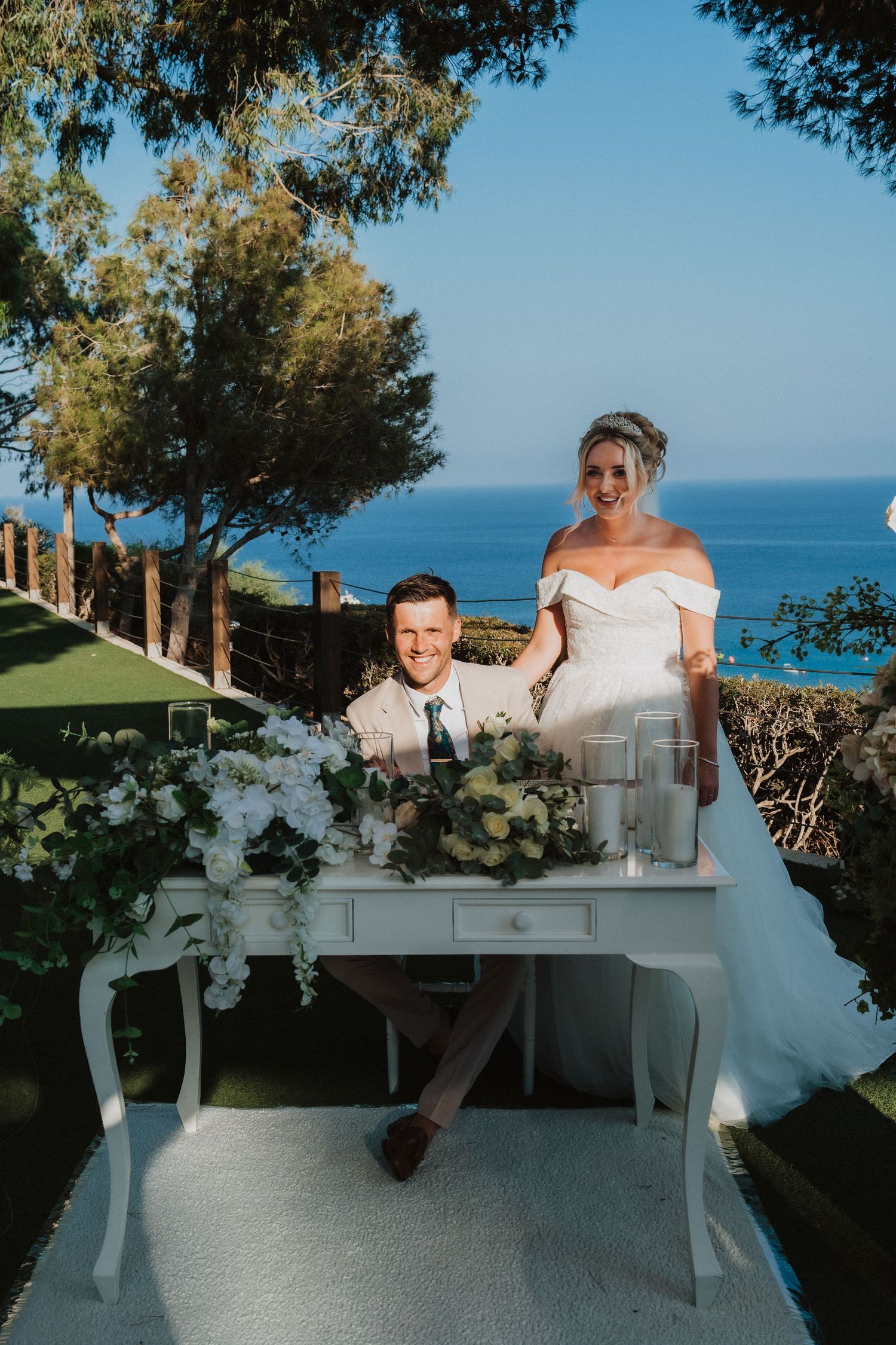 A couple at their outdoor wedding ceremony with a scenic coastal view and white desk decorated with flowers.