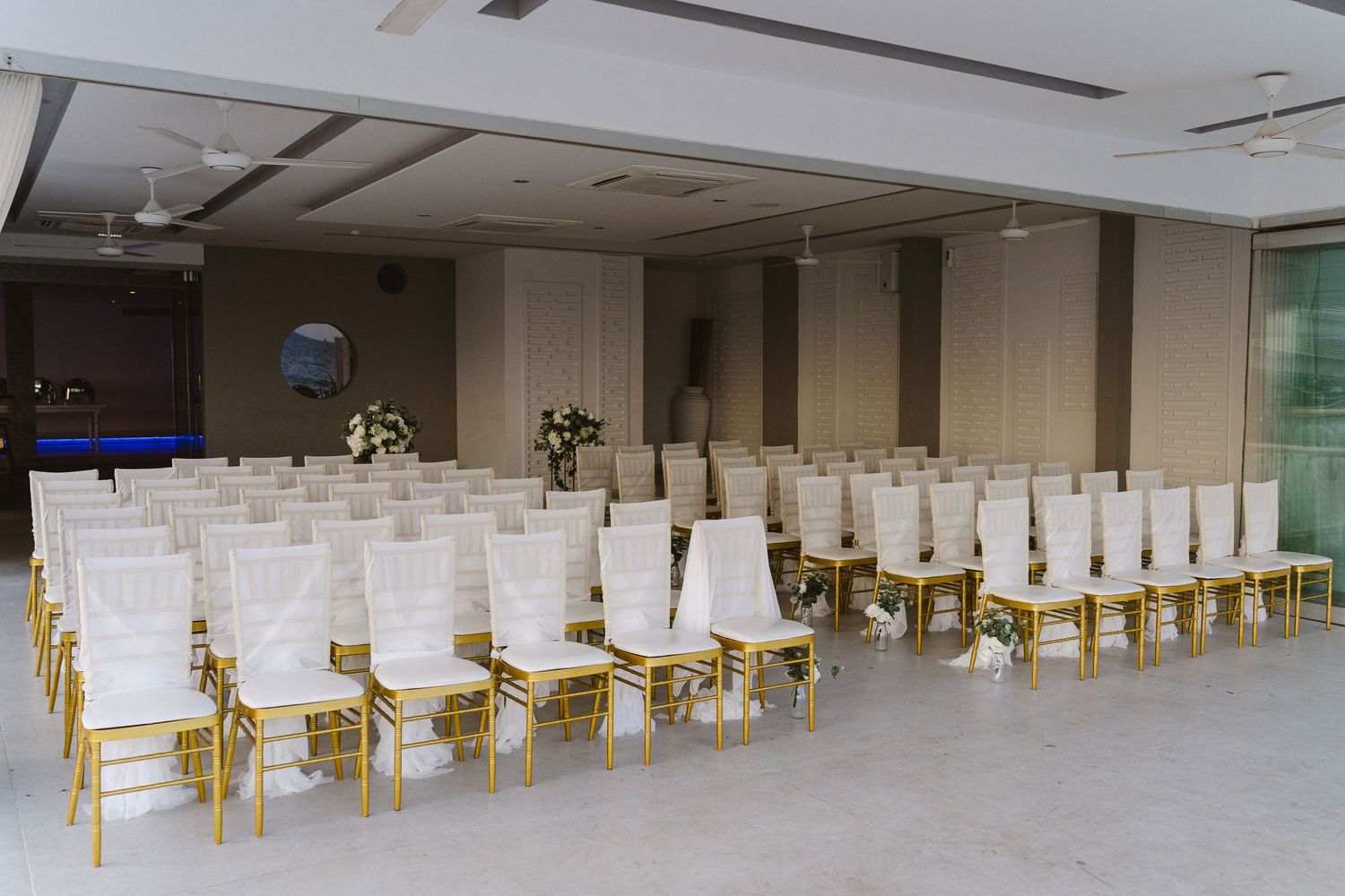 Modern indoor venue setup with rows of white-covered chairs and gold frames for a ceremony.