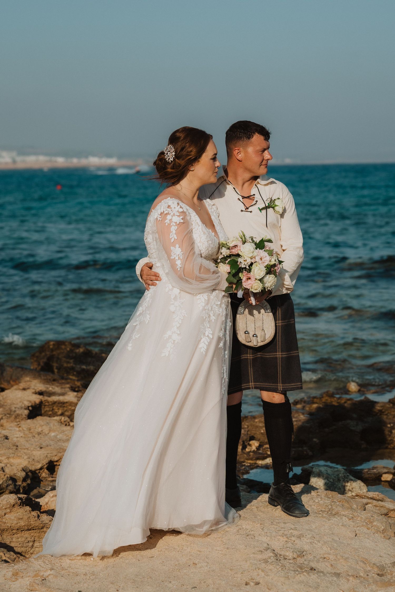 A coastal wedding portrait featuring elegant bridal attire and Scottish formal wear on rocky shores.
