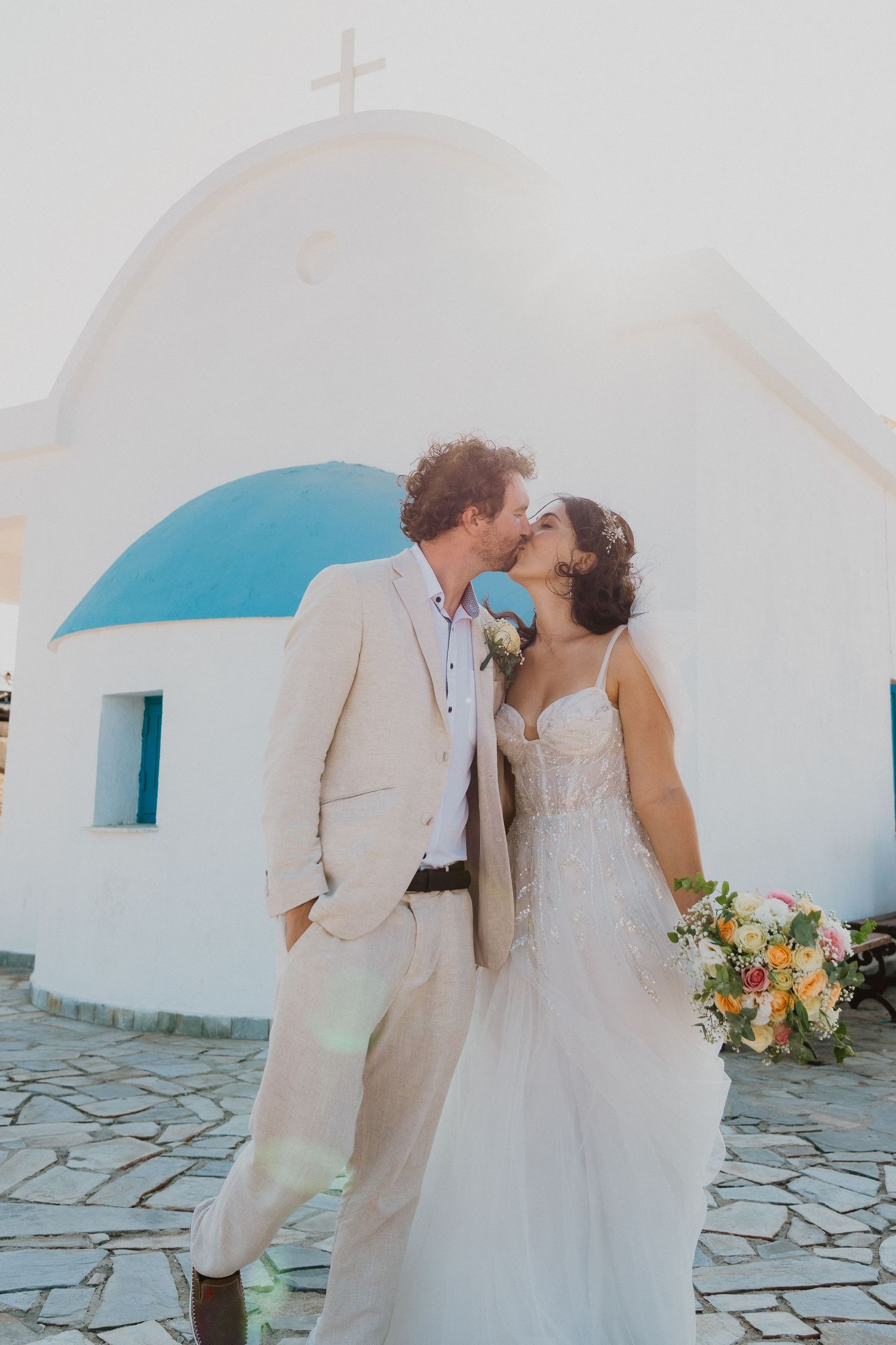 Newlyweds share romantic moment in front of white Greek church with blue dome on warm summer day.