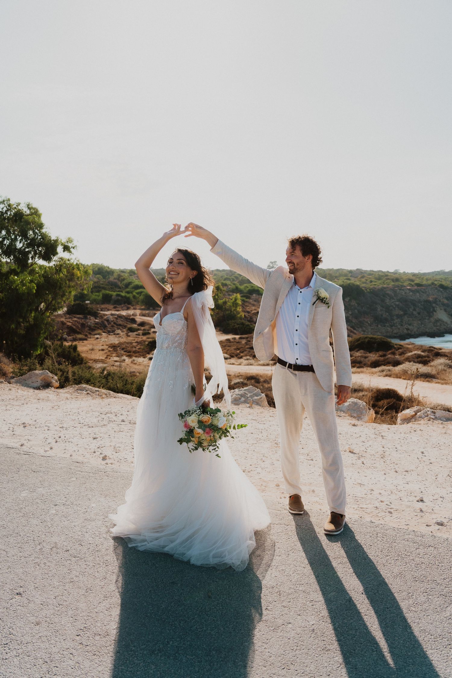 A couple in wedding attire dance together on a scenic overlook with natural rock formations and greenery in the background.