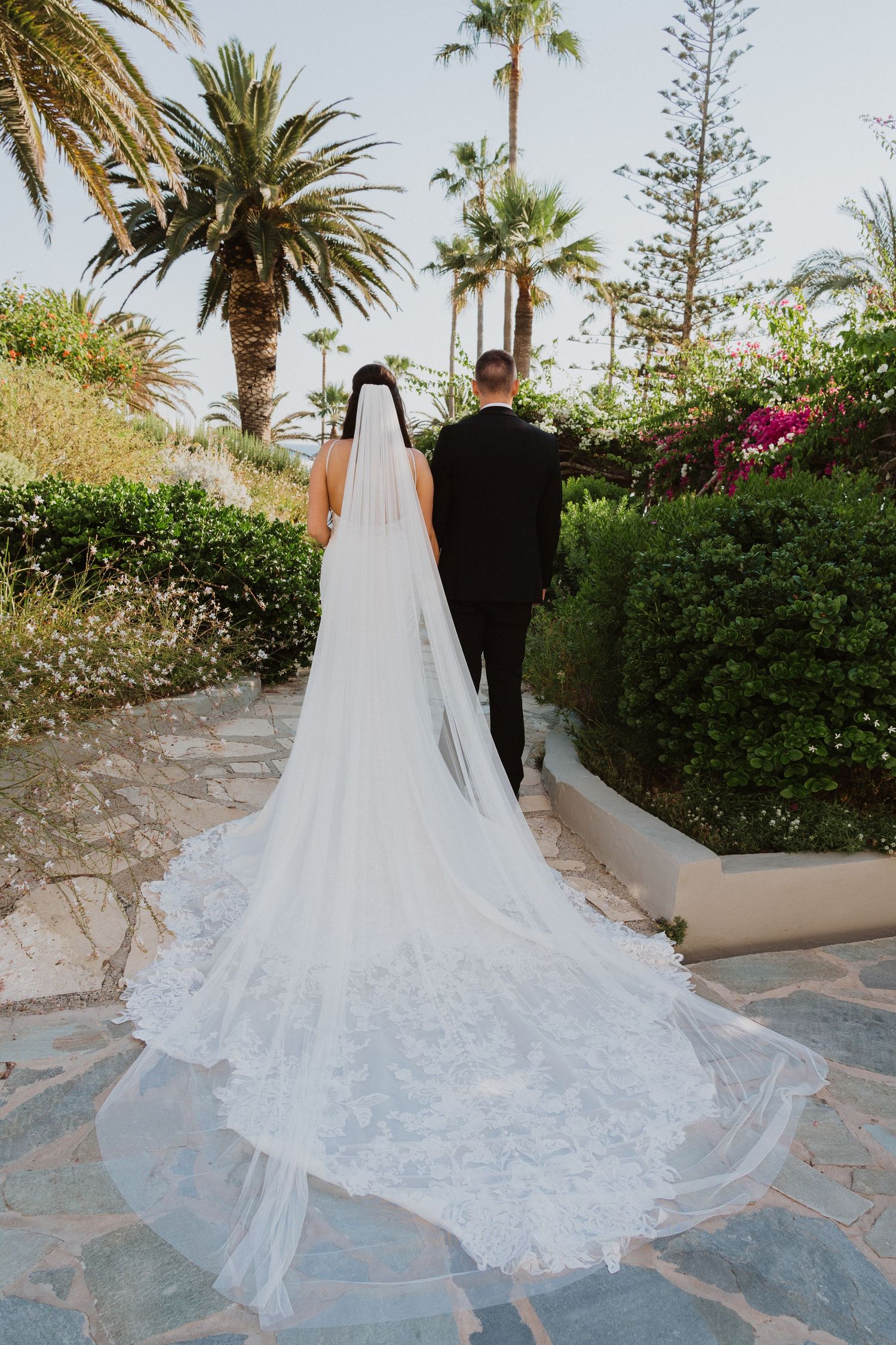 A flowing white wedding dress train trails across a stone pathway surrounded by palm trees.
