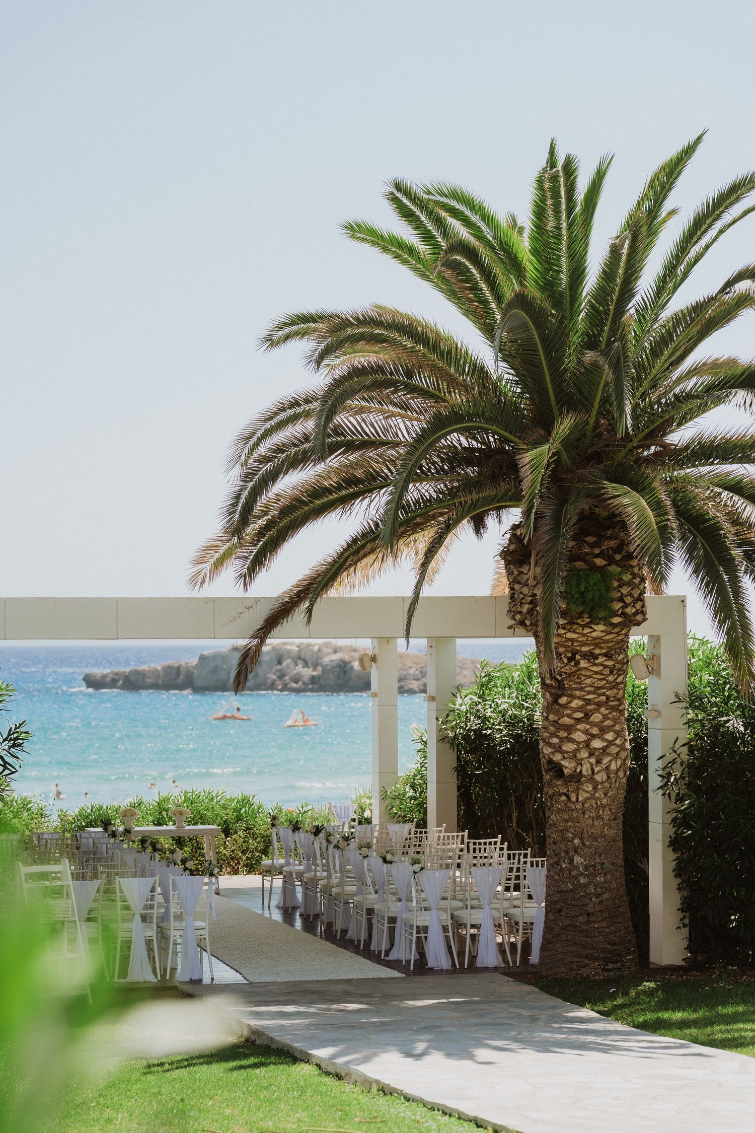 Palm trees line an elegant outdoor wedding venue with white chairs overlooking a tropical beach and ocean view.