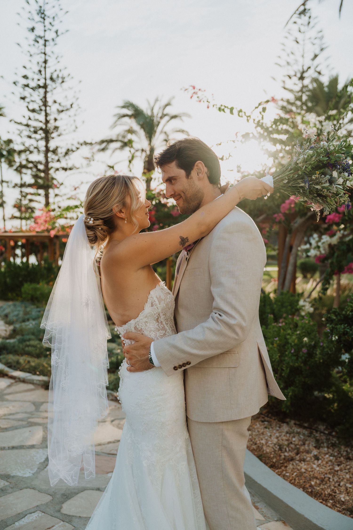Wedding couple embraces on a garden path during golden hour with palm trees in the background.
