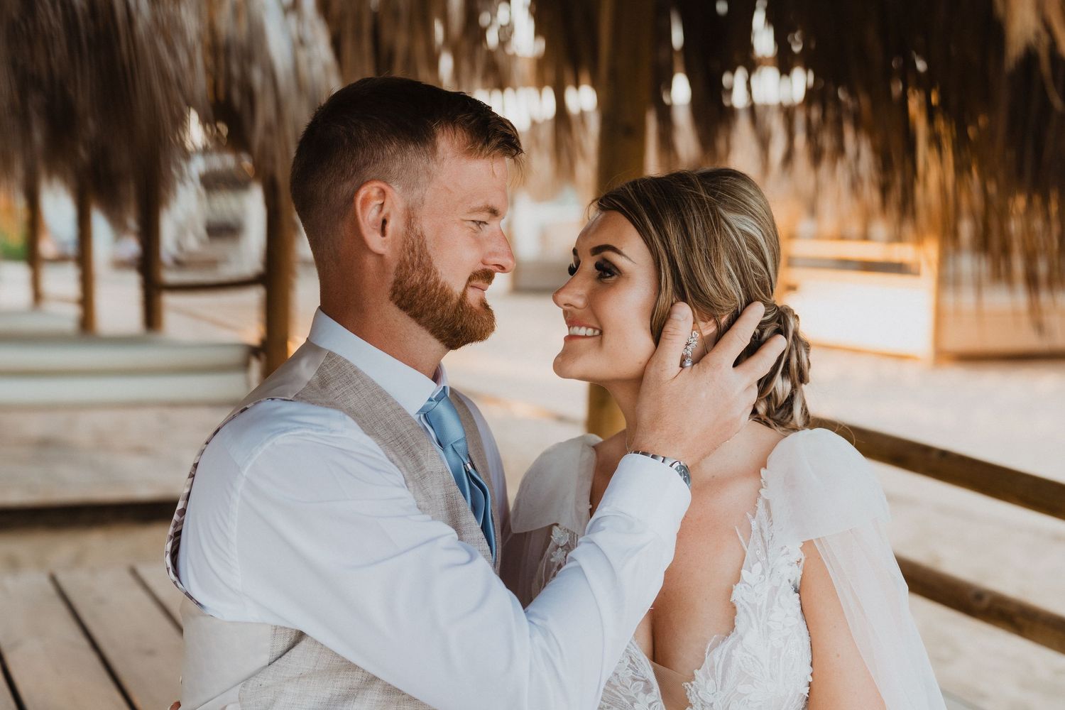 Wedding couple shares tender moments during beach photoshoot under thatched shelter.