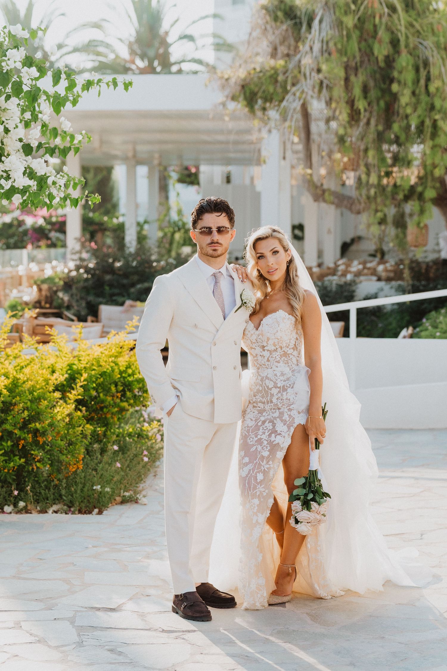 A couple in white wedding attire walking together on a bright Mediterranean-style patio with lush gardens.