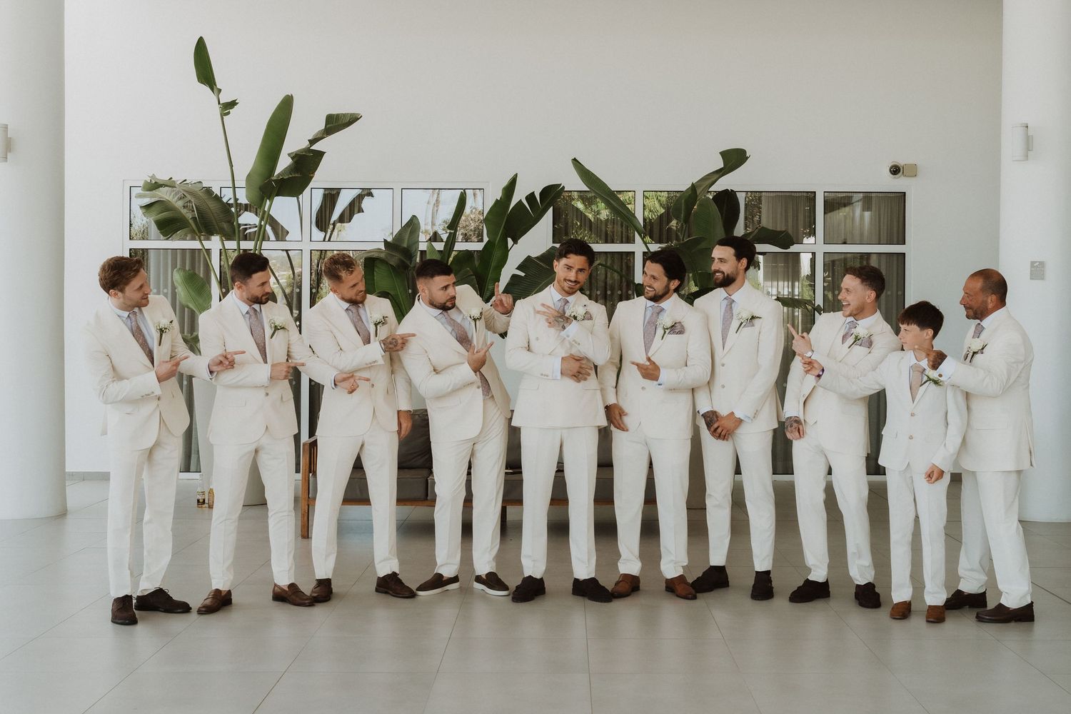 A group of groomsmen in all-white suits stand in a line against a white wall at a modern wedding venue.