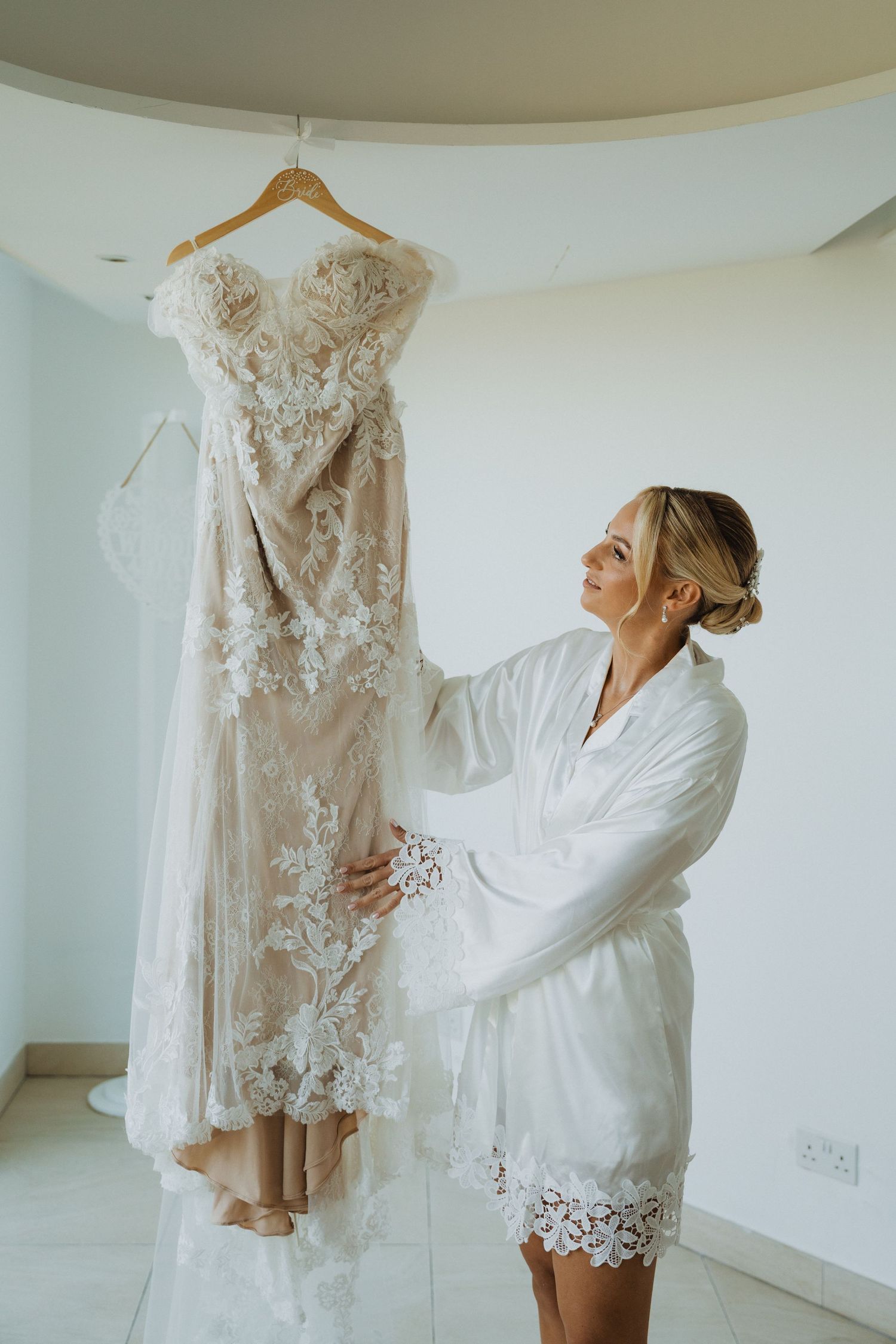 Elegant lace wedding dress hanging while being admired in bright white room.