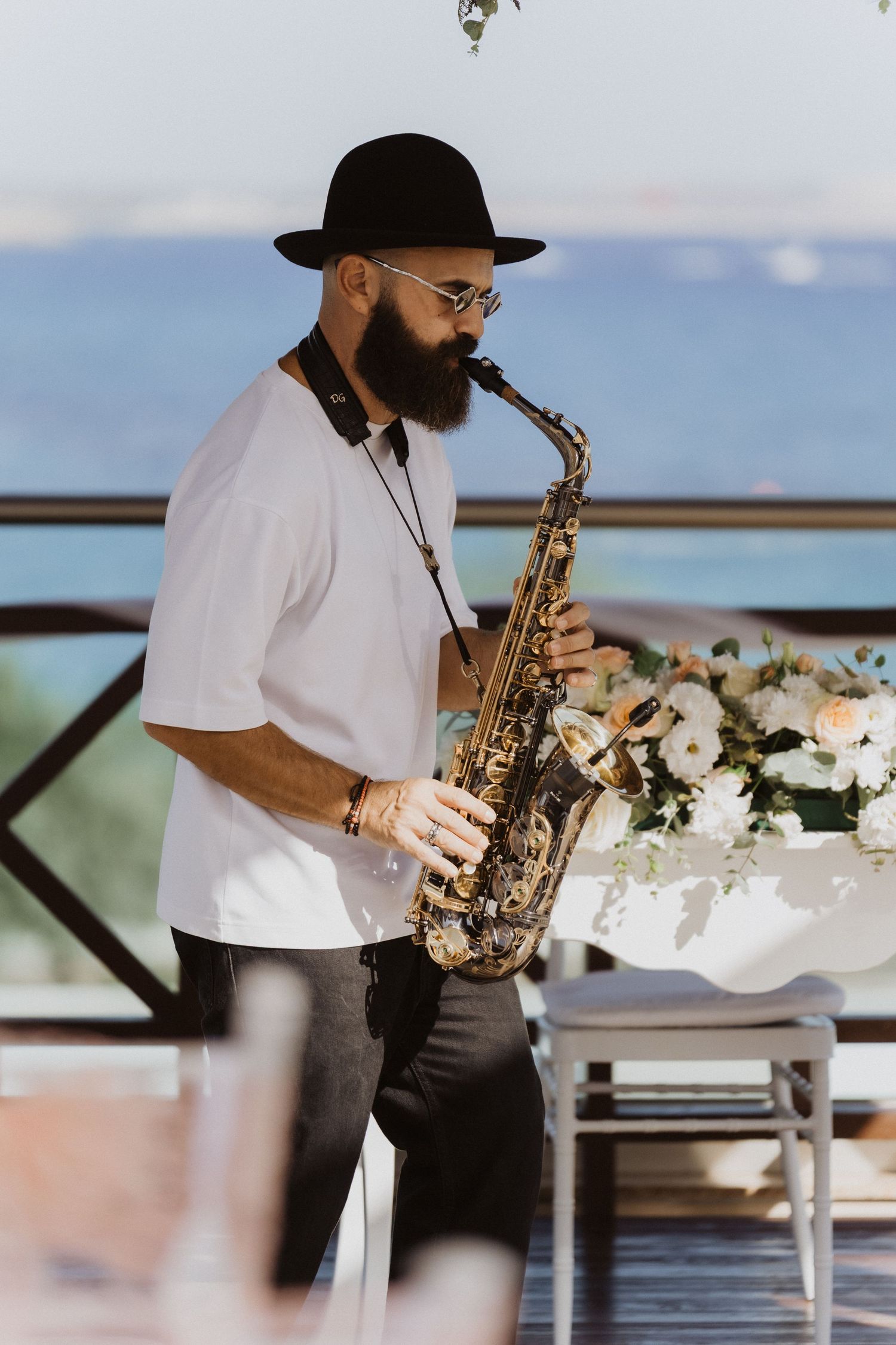 Bearded musician playing saxophone at outdoor venue with ocean view and white flowers.