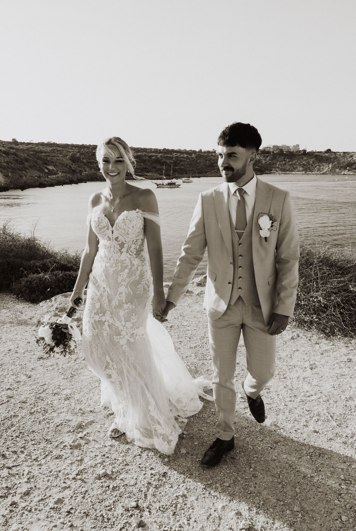 Black and white wedding photo of couple walking along scenic coastal path during sunset.