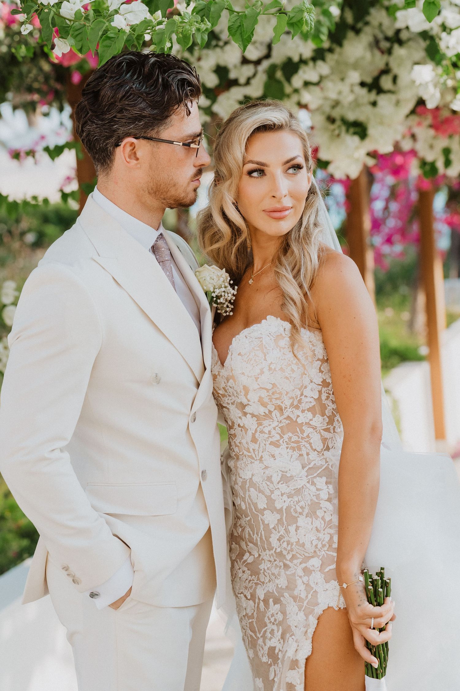 Elegant wedding couple in cream and white attire pose together outdoors with pink bougainvillea flowers in background.