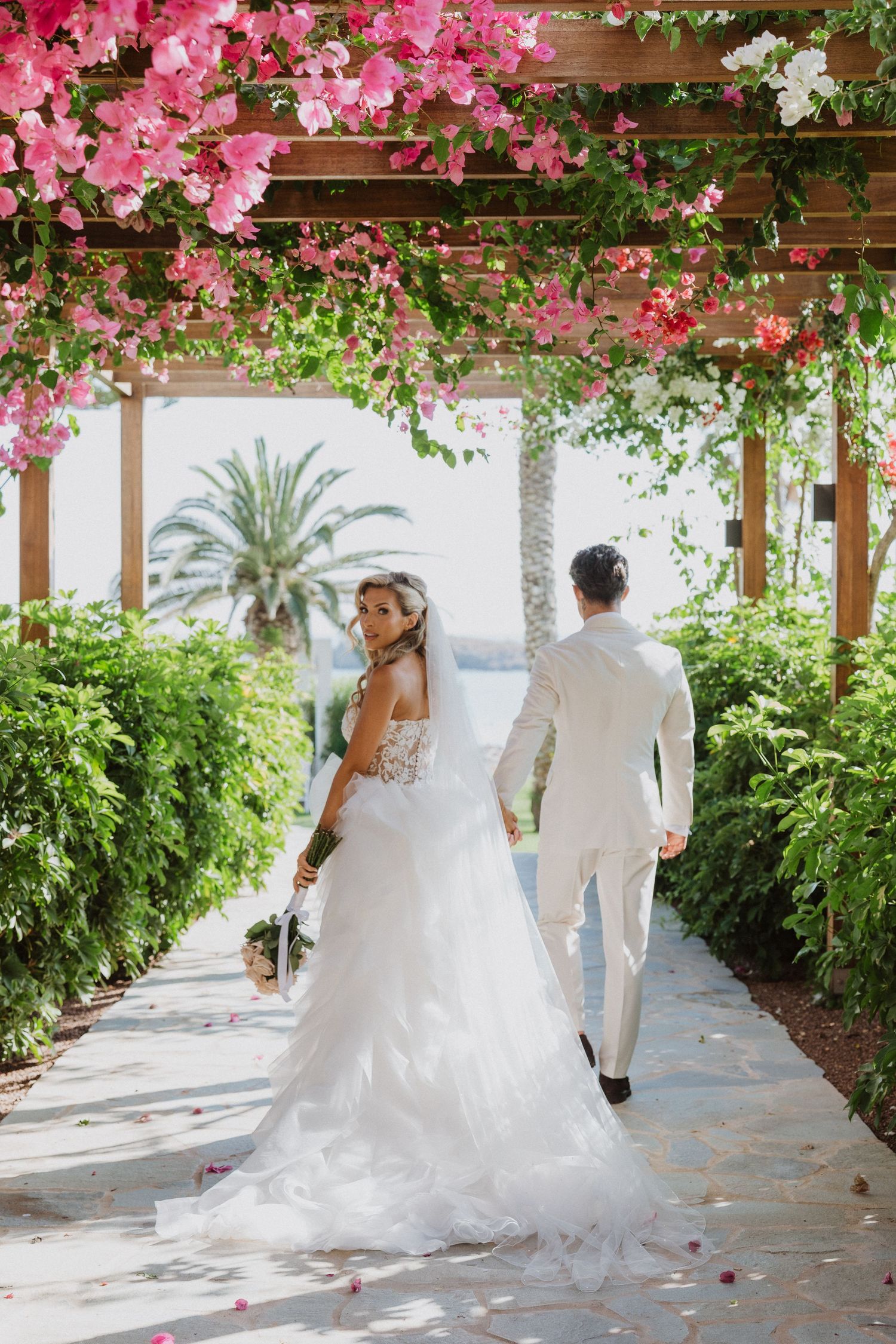 A couple walks under a pergola covered in pink bougainvillea flowers at a tropical wedding venue.