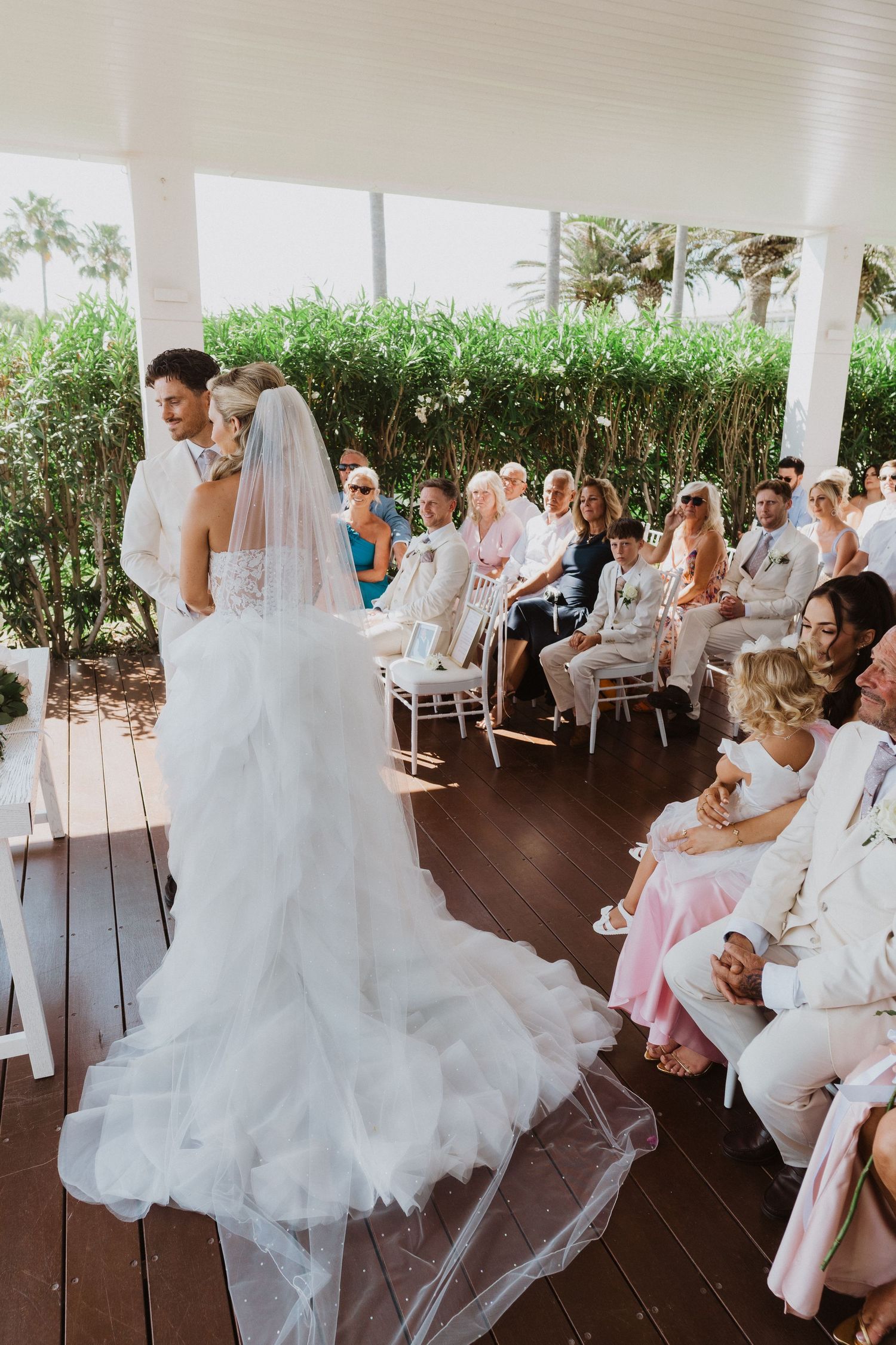 A wedding ceremony takes place on a covered deck with guests seated in white chairs and greenery in the background.