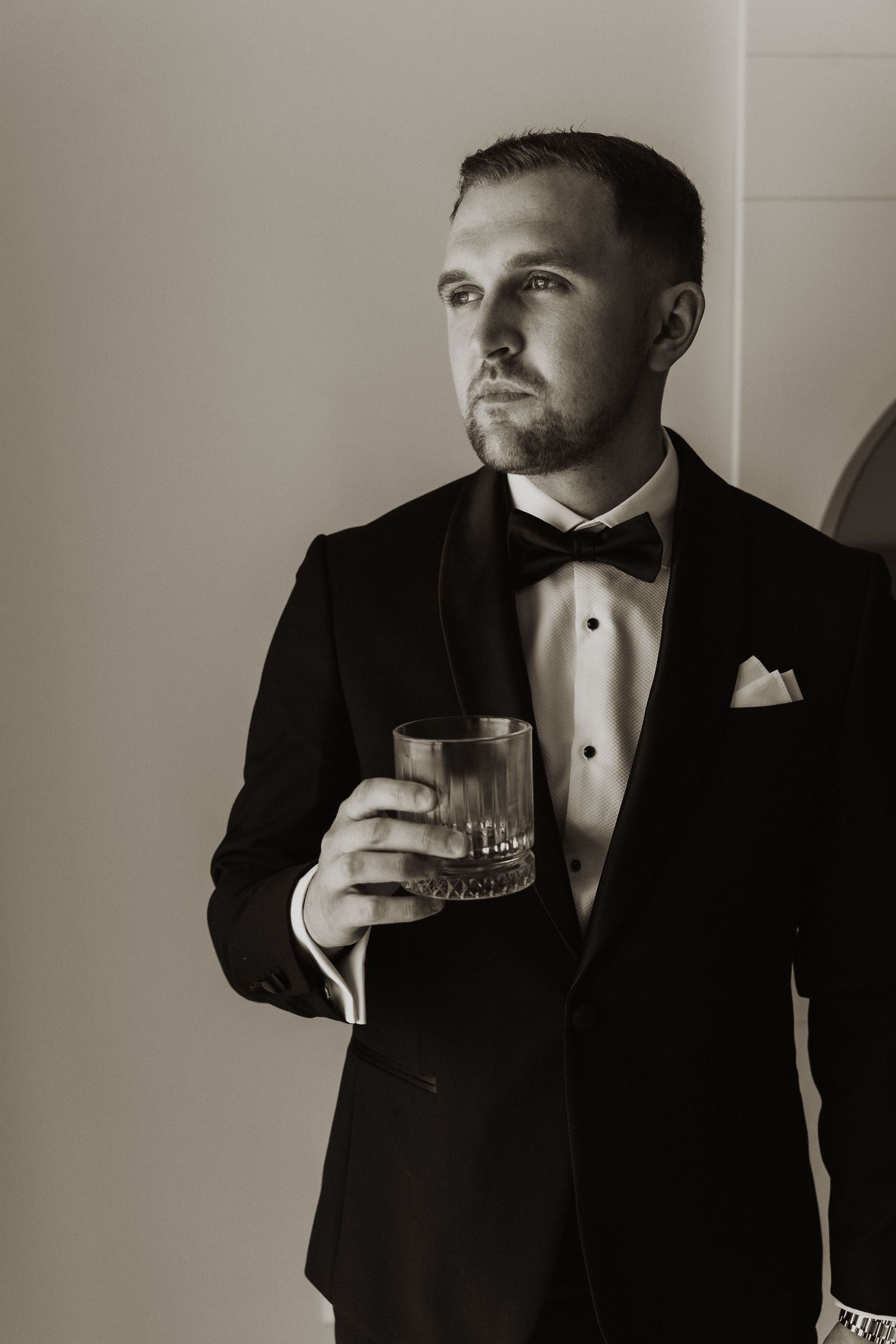 Elegant groom in black tuxedo with bow tie holding whiskey glass in moody black and white portrait.