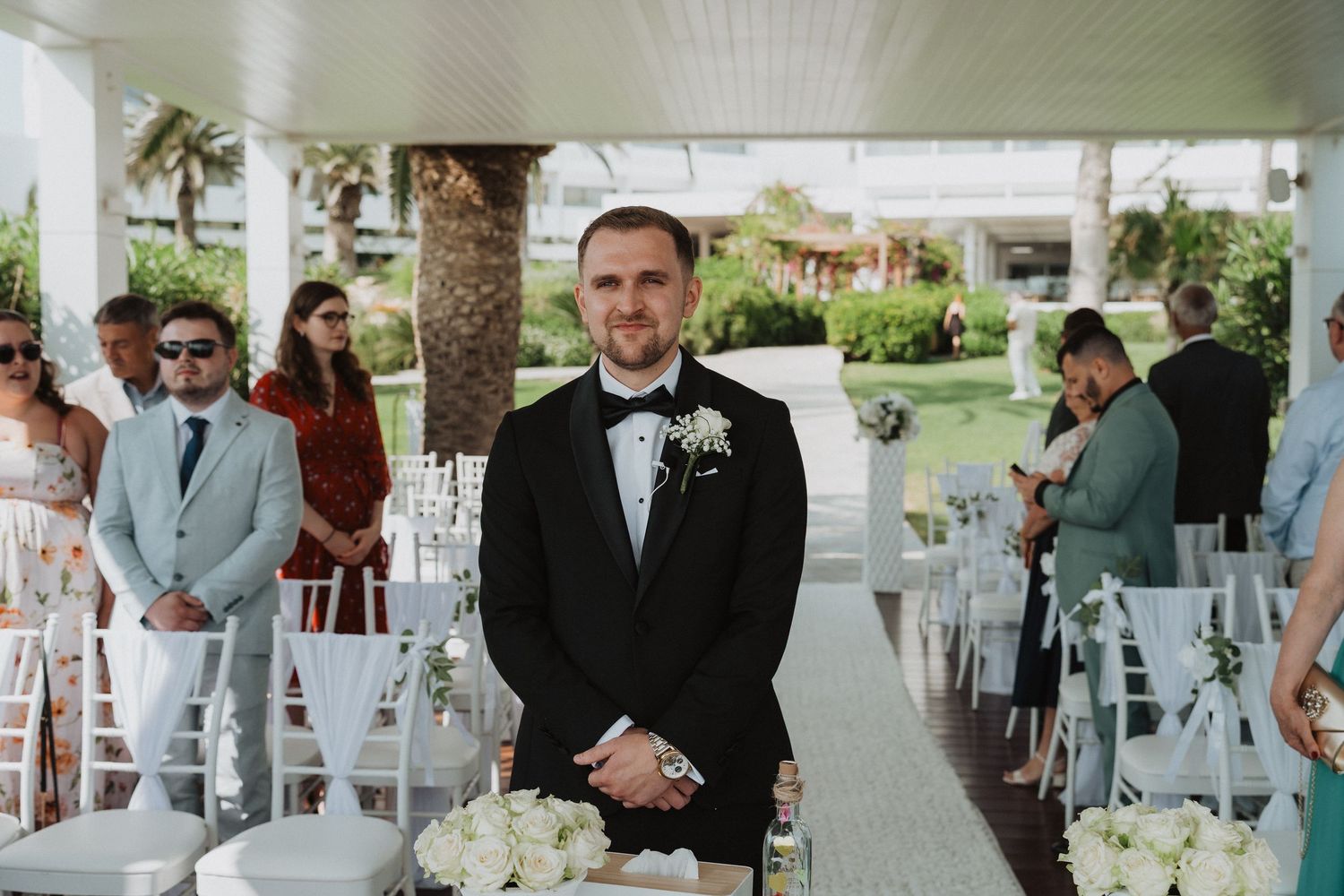 Groom in black tuxedo standing at outdoor wedding ceremony aisle with white floral arrangements.