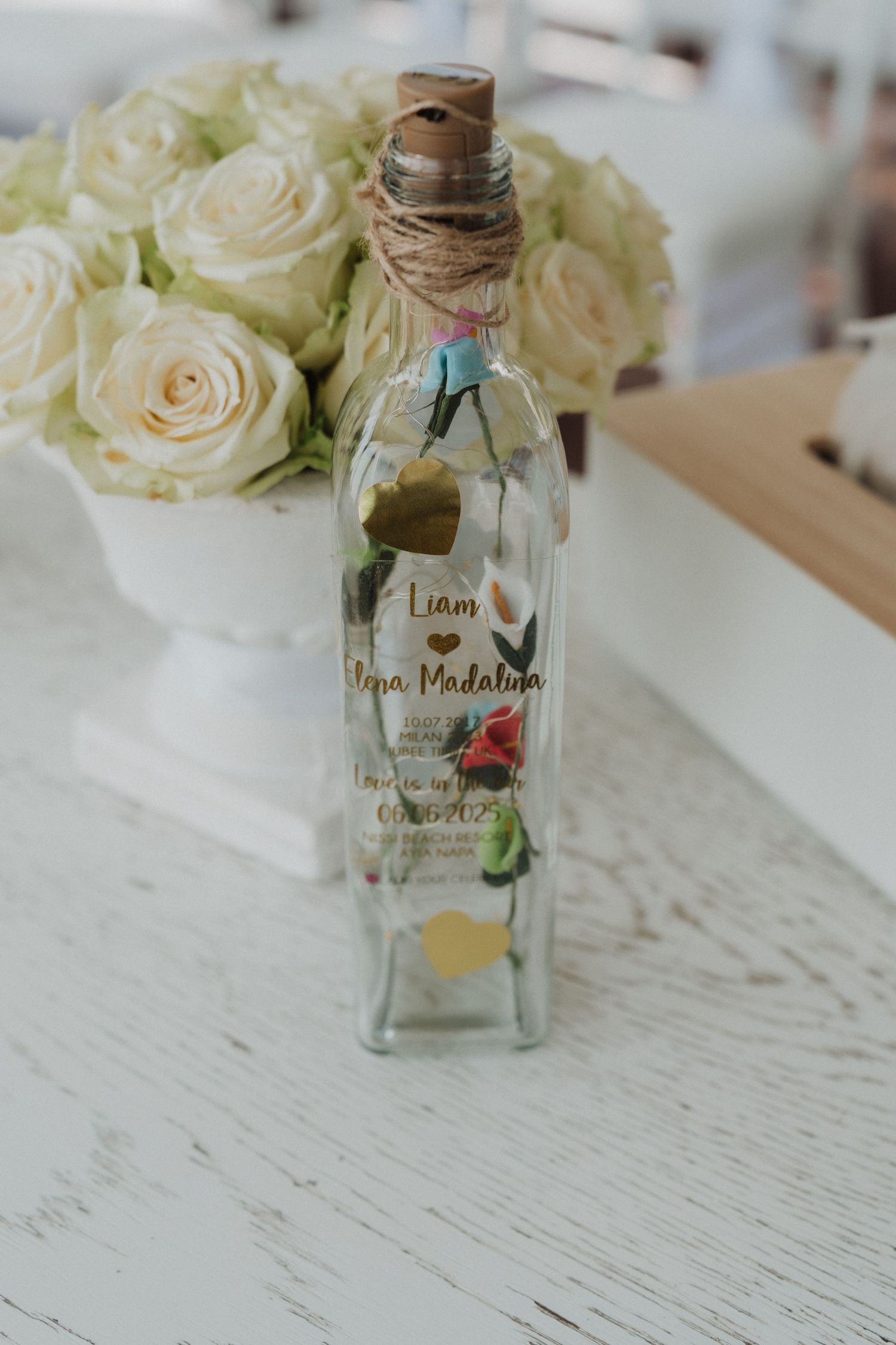A decorative glass bottle with twine stopper inscribed with wedding names Liam and Ana Madalina beside white roses.