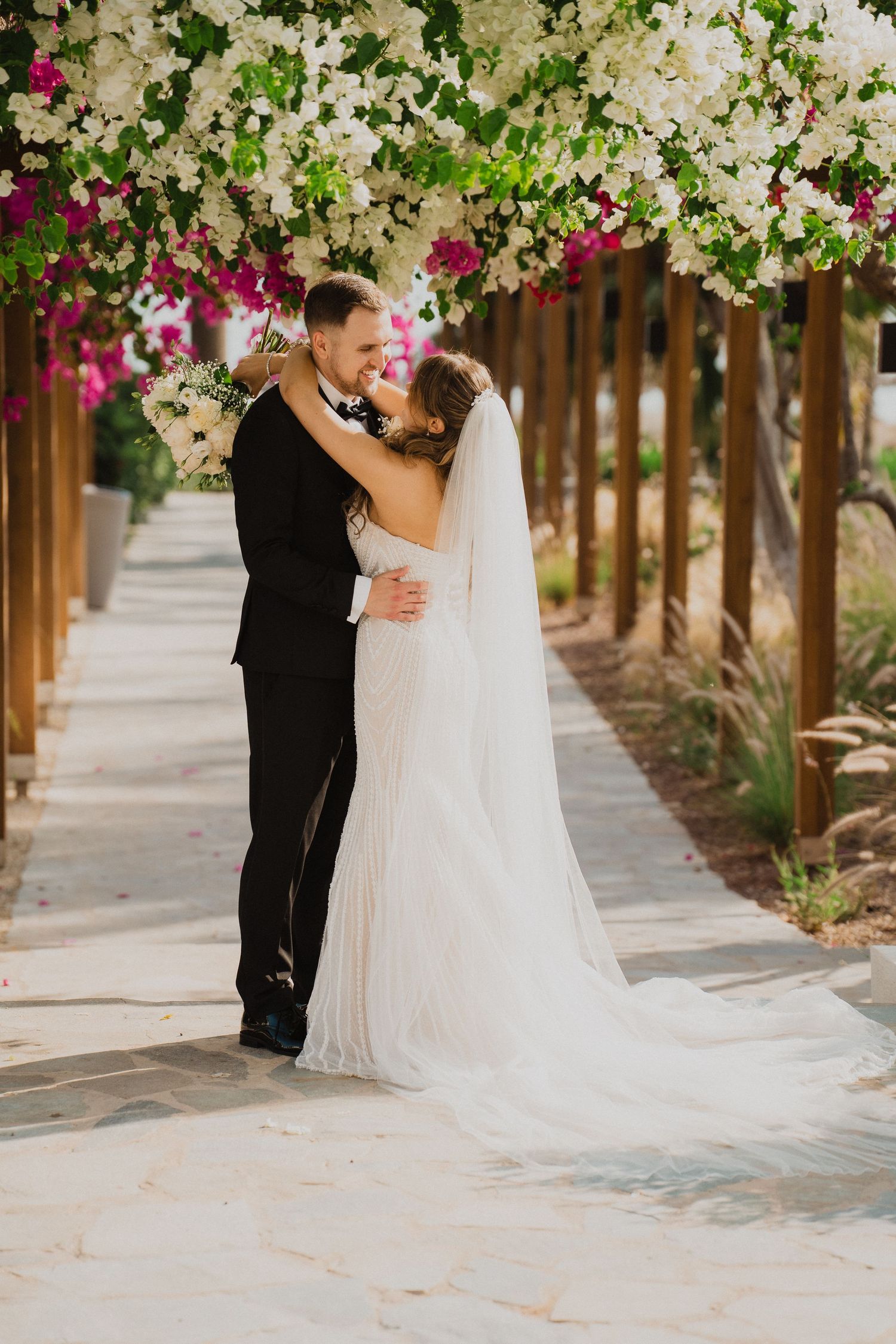 Bride and groom embracing under a floral arch of white and pink bougainvillea on a stone pathway.