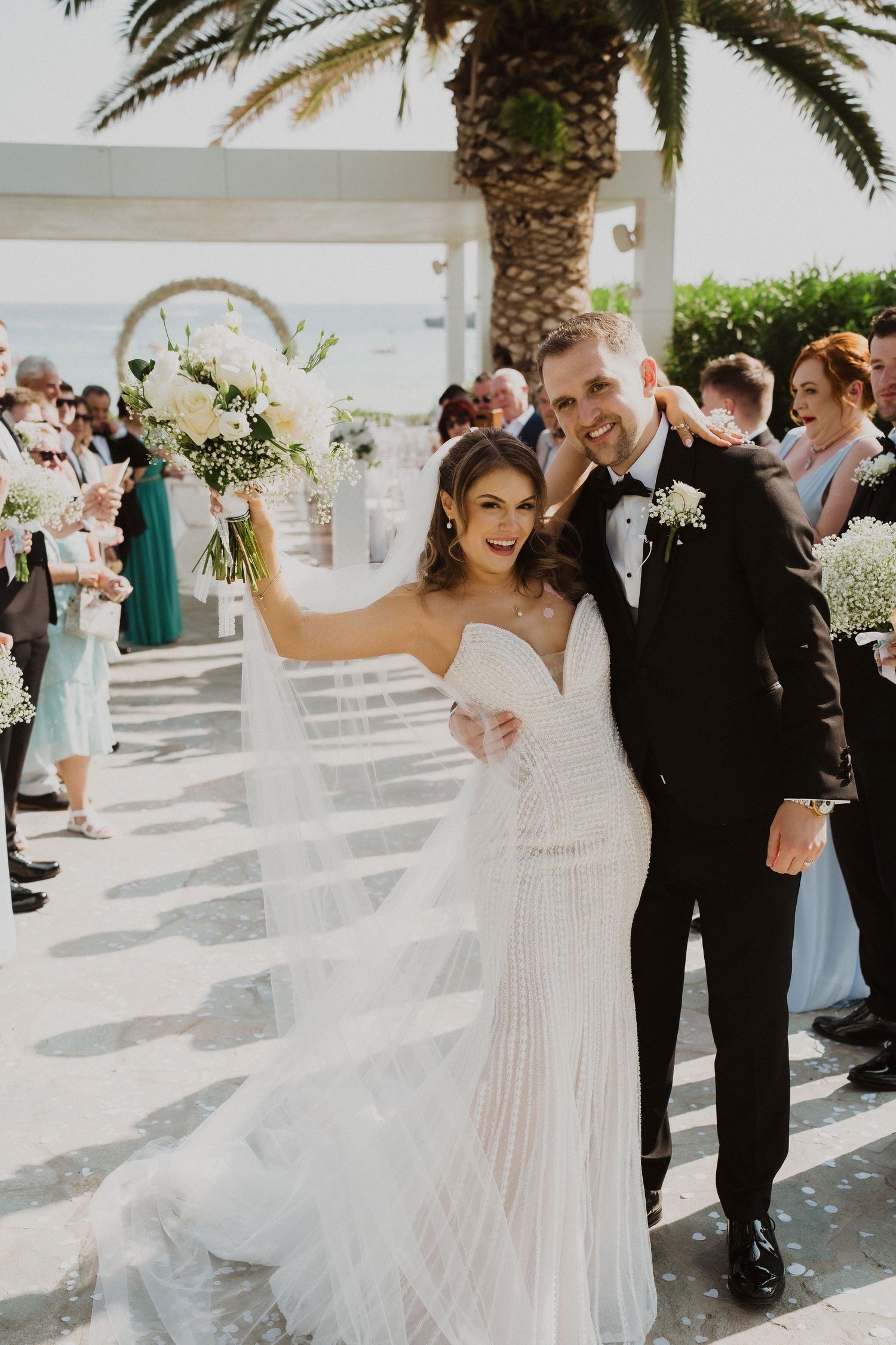Radiant bride in beaded gown raises bouquet beside groom in black tuxedo after outdoor beach wedding ceremony.
