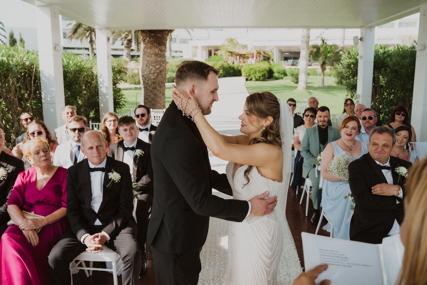 Bride and groom sharing first kiss at altar as wedding guests watch during outdoor ceremony.