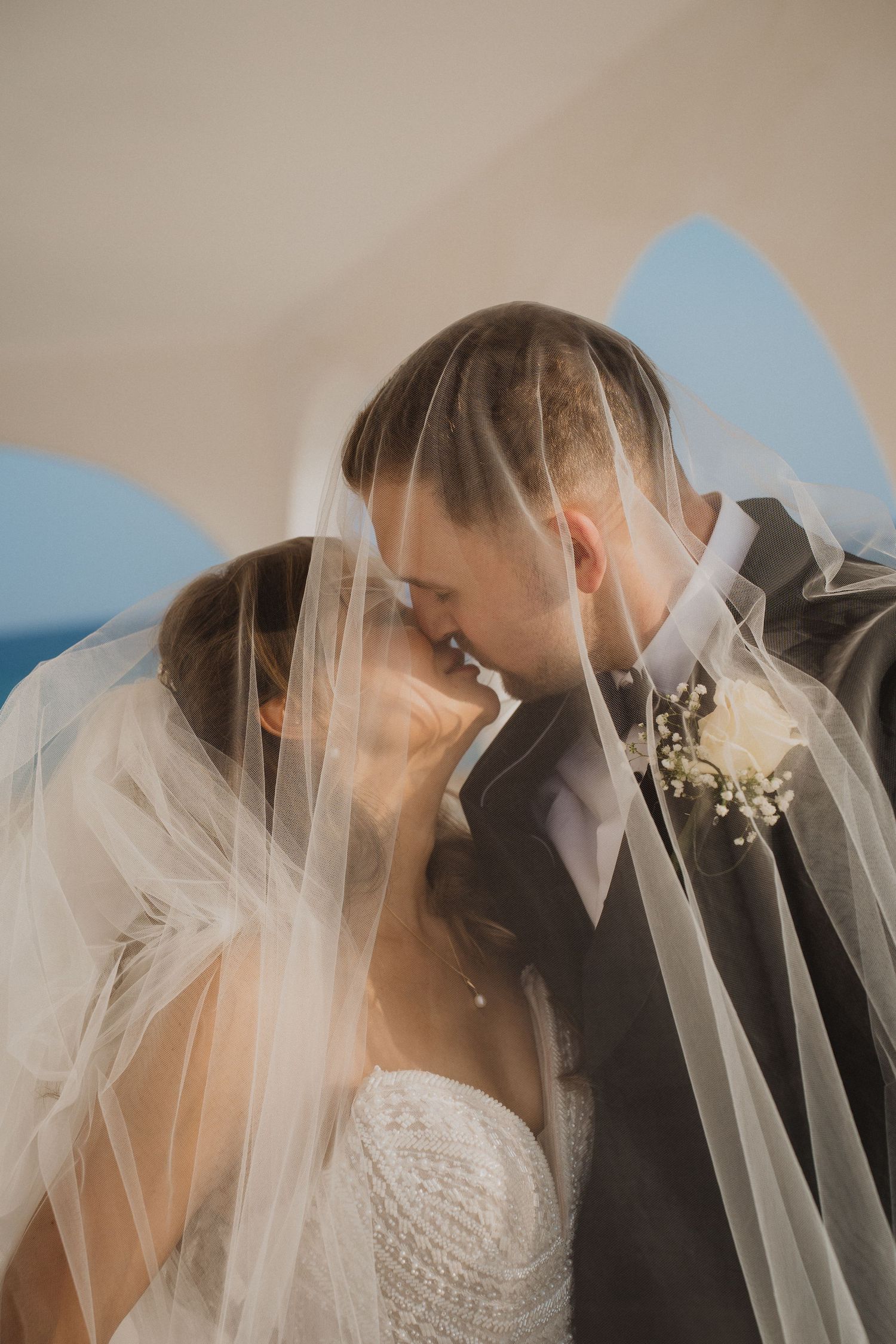 Romantic bride and groom sharing a kiss beneath flowing white wedding veil with ocean backdrop at coastal wedding.