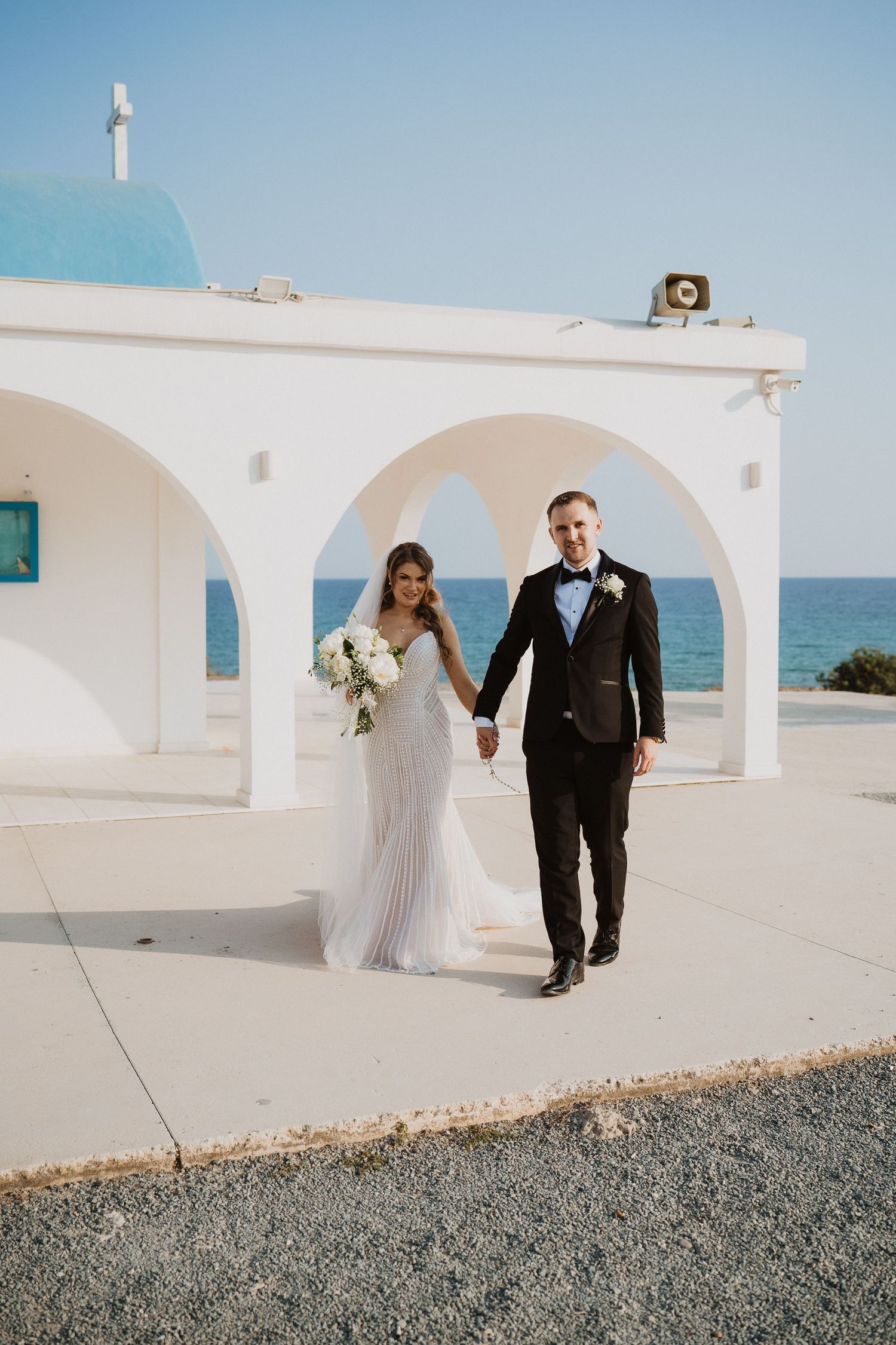 Newlyweds hold hands walking past a white seaside chapel with blue dome on their wedding day.