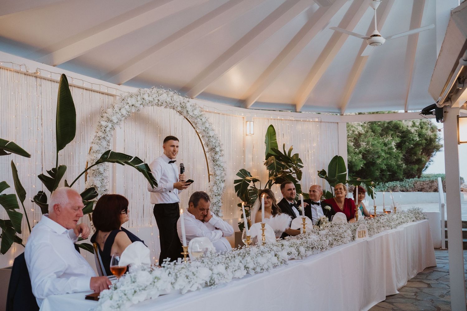 Wedding guests seated at a long floral table under a marquee during a elegant reception speech.