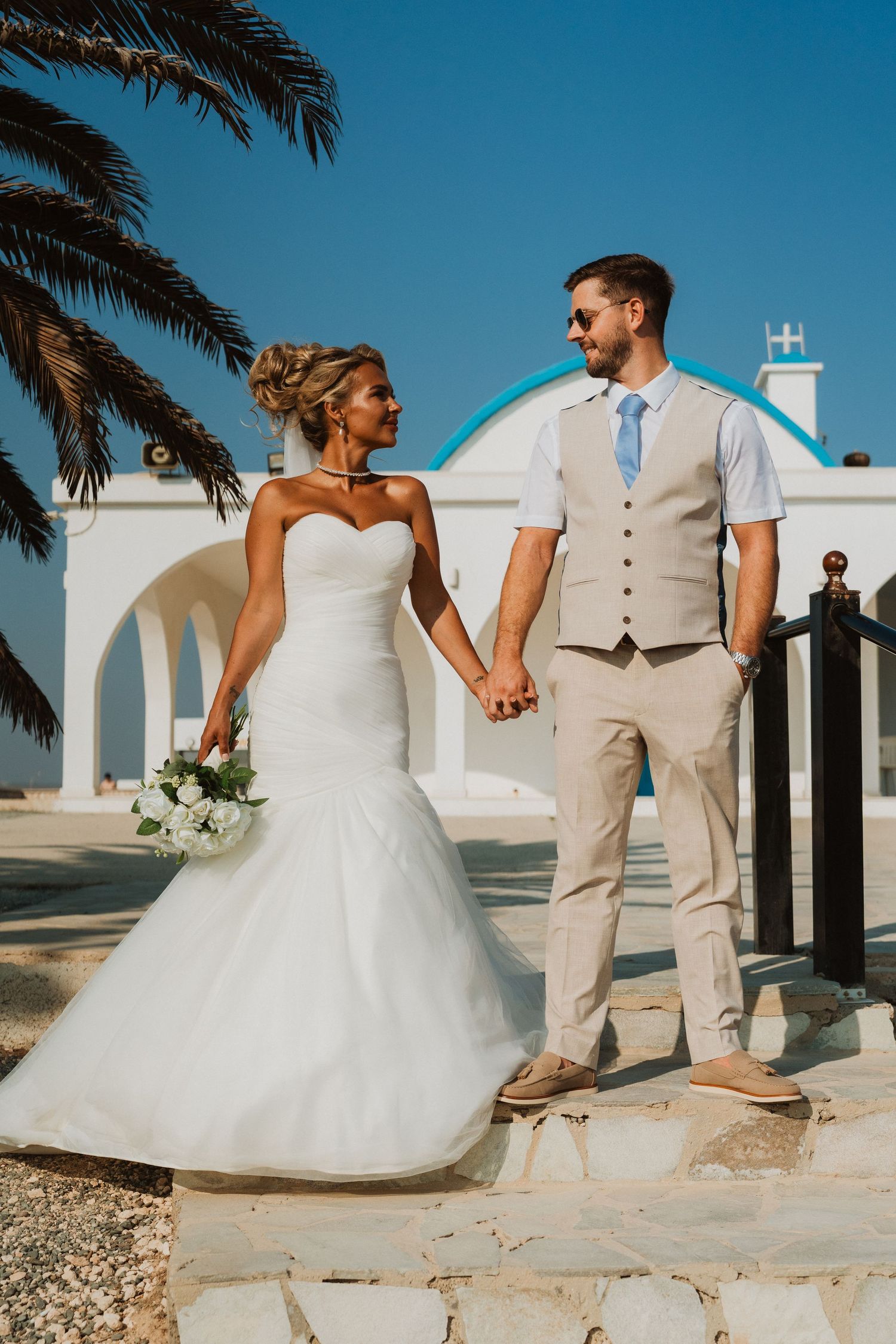 Bride and groom hold hands outside a white Greek chapel with blue dome, palm tree, and clear blue sky.