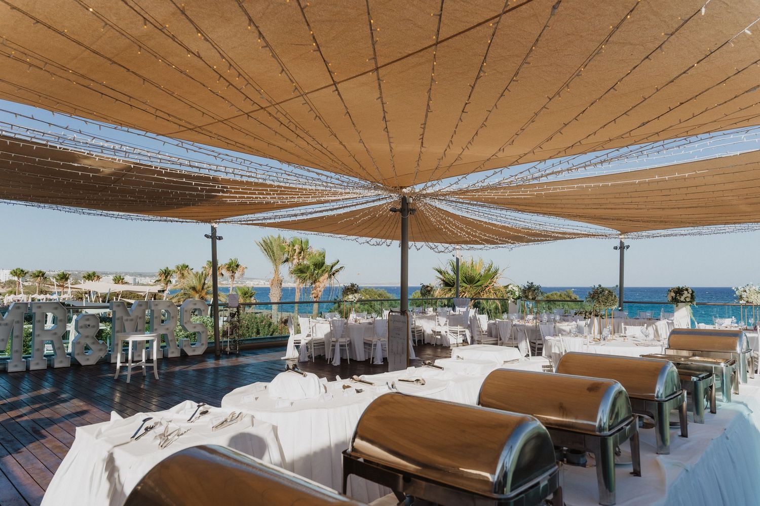 Beachfront restaurant terrace with tan awnings, white tablecloths and comfortable seating overlooking the ocean.