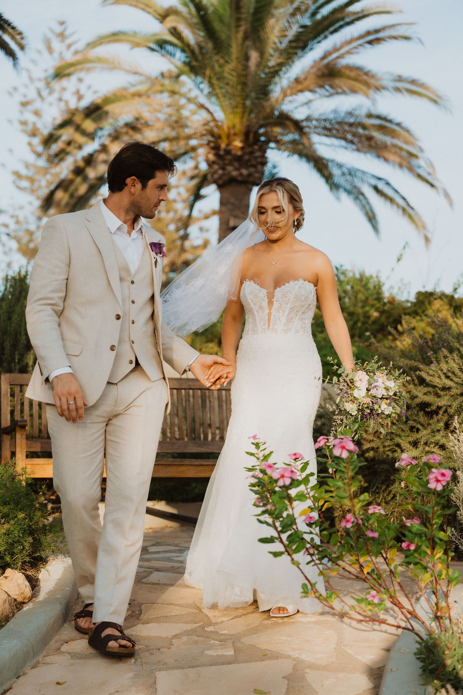 Newlyweds in a tropical garden setting with palm trees, bride in white gown and groom in light beige suit.