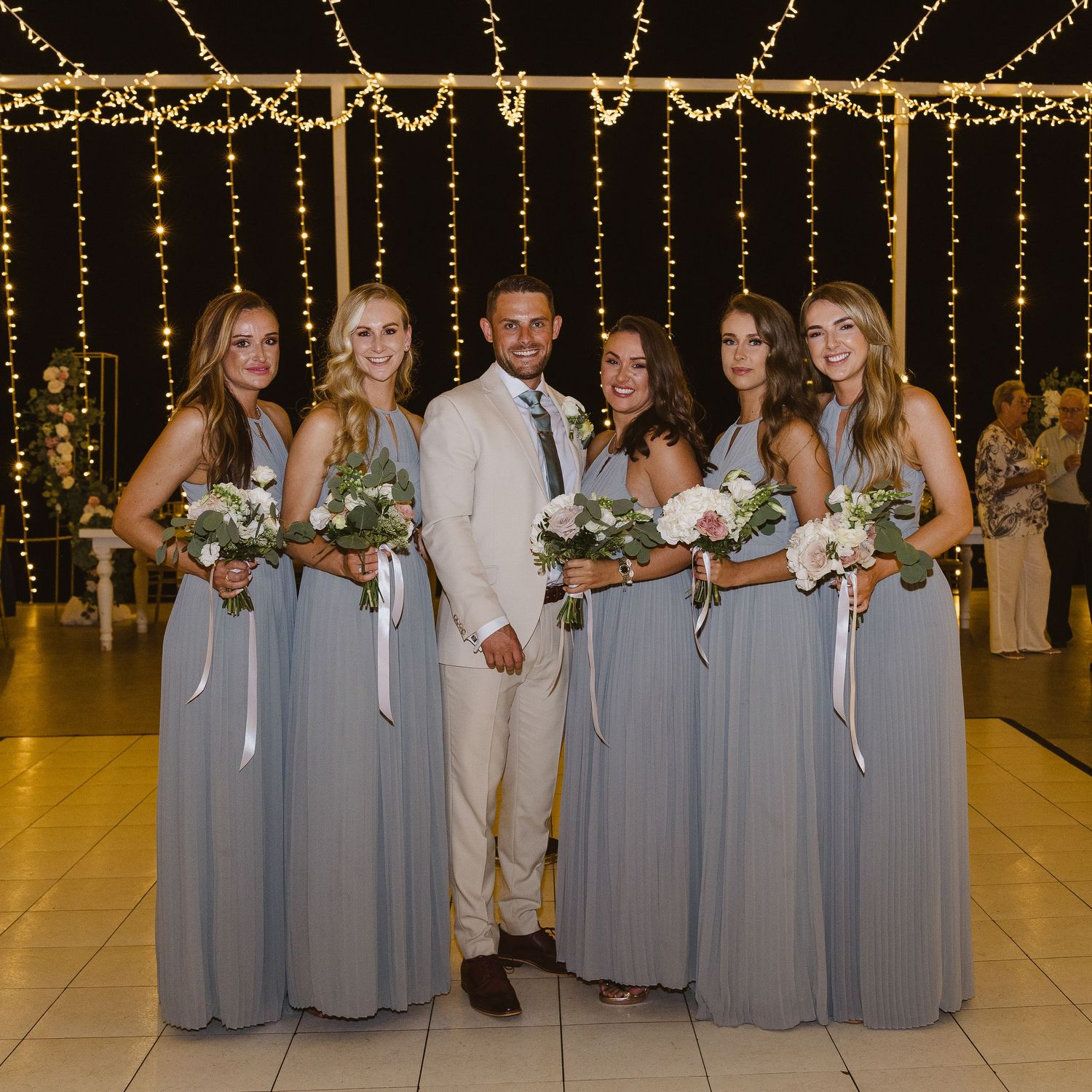 Wedding party in grey dresses and beige suit poses under warm string lights.