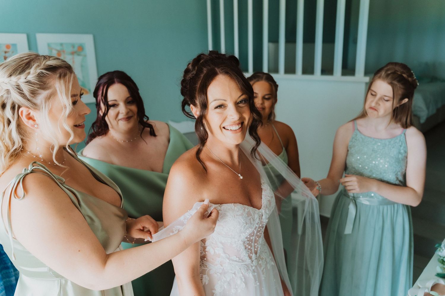 Joyful bridesmaids in mint green dresses help the bride prepare before a wedding ceremony.