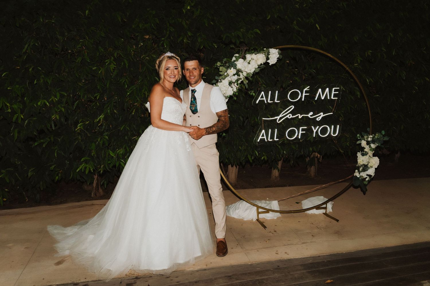 Wedding ceremony backdrop with illuminated arch and 'All of Me All of You' sign decorated with flowers.