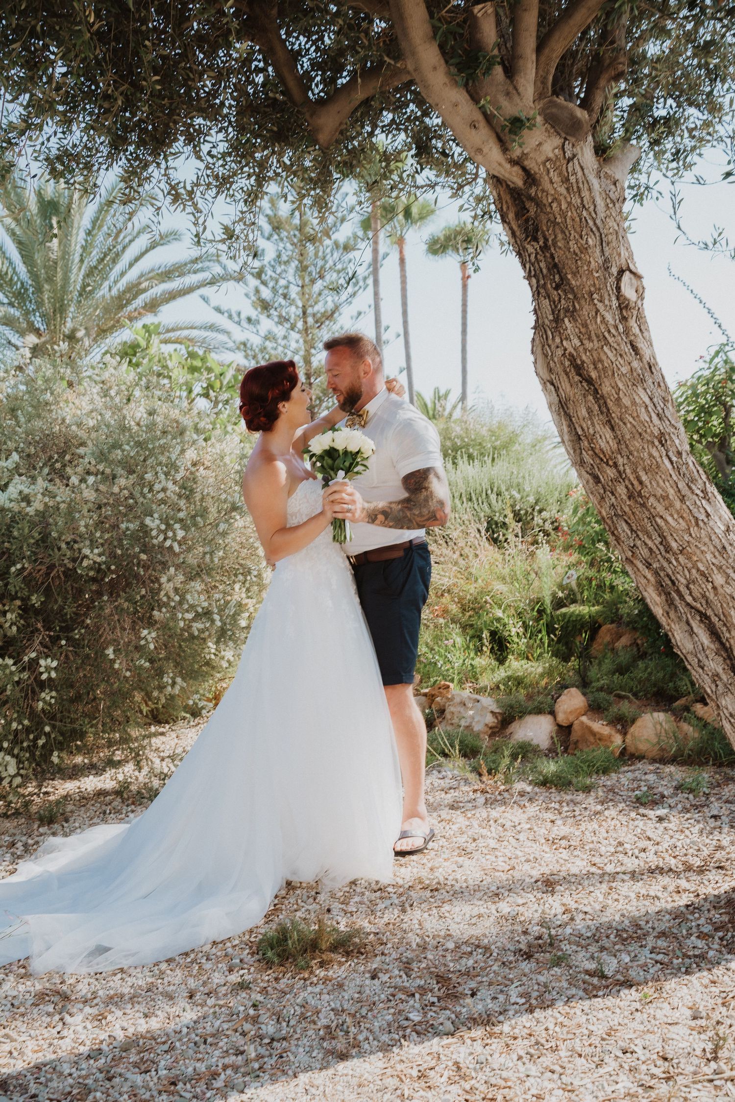 Newlyweds share intimate moment under large tree in tropical garden setting.