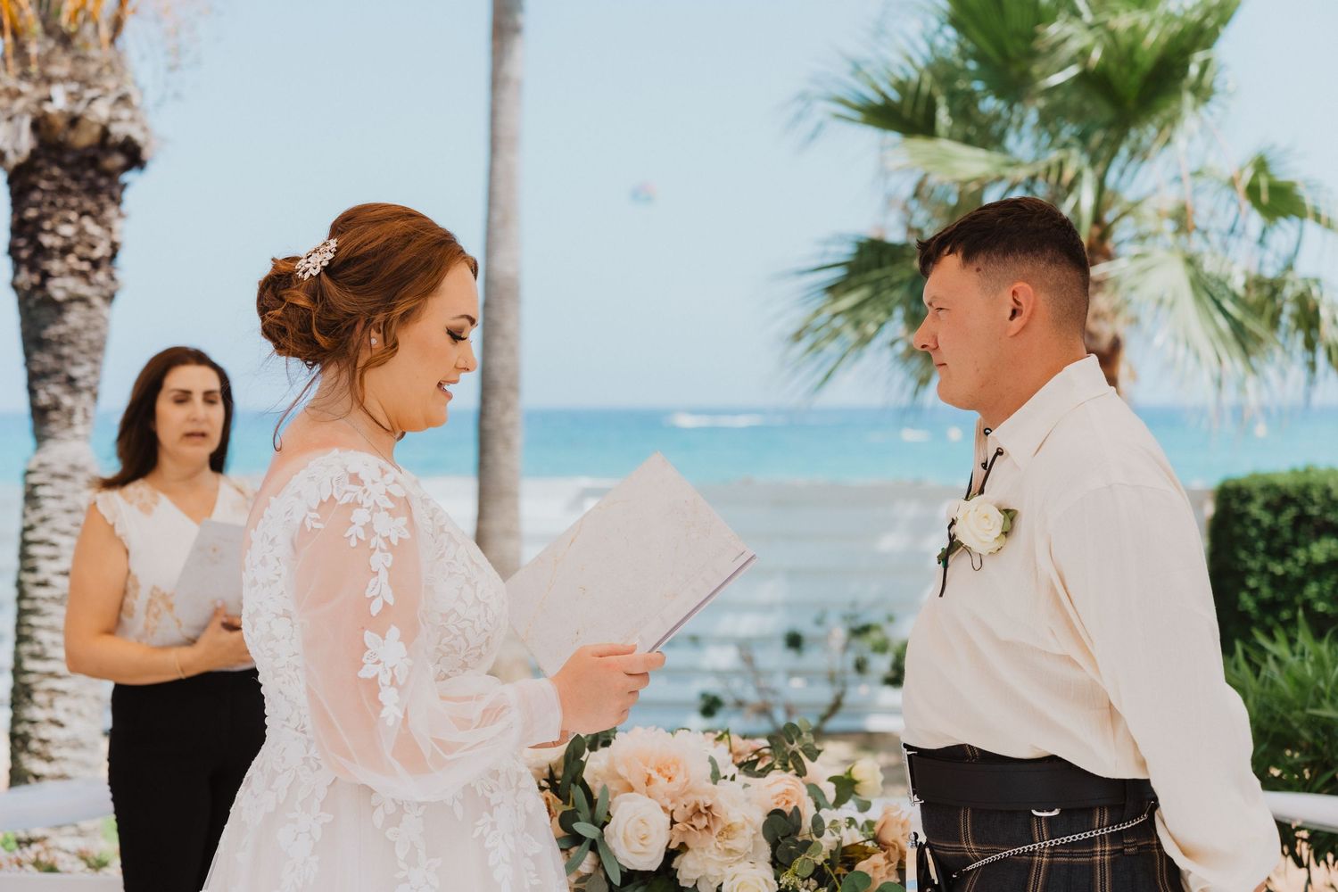 A beautiful outdoor wedding ceremony takes place by the ocean with palm trees in the background.