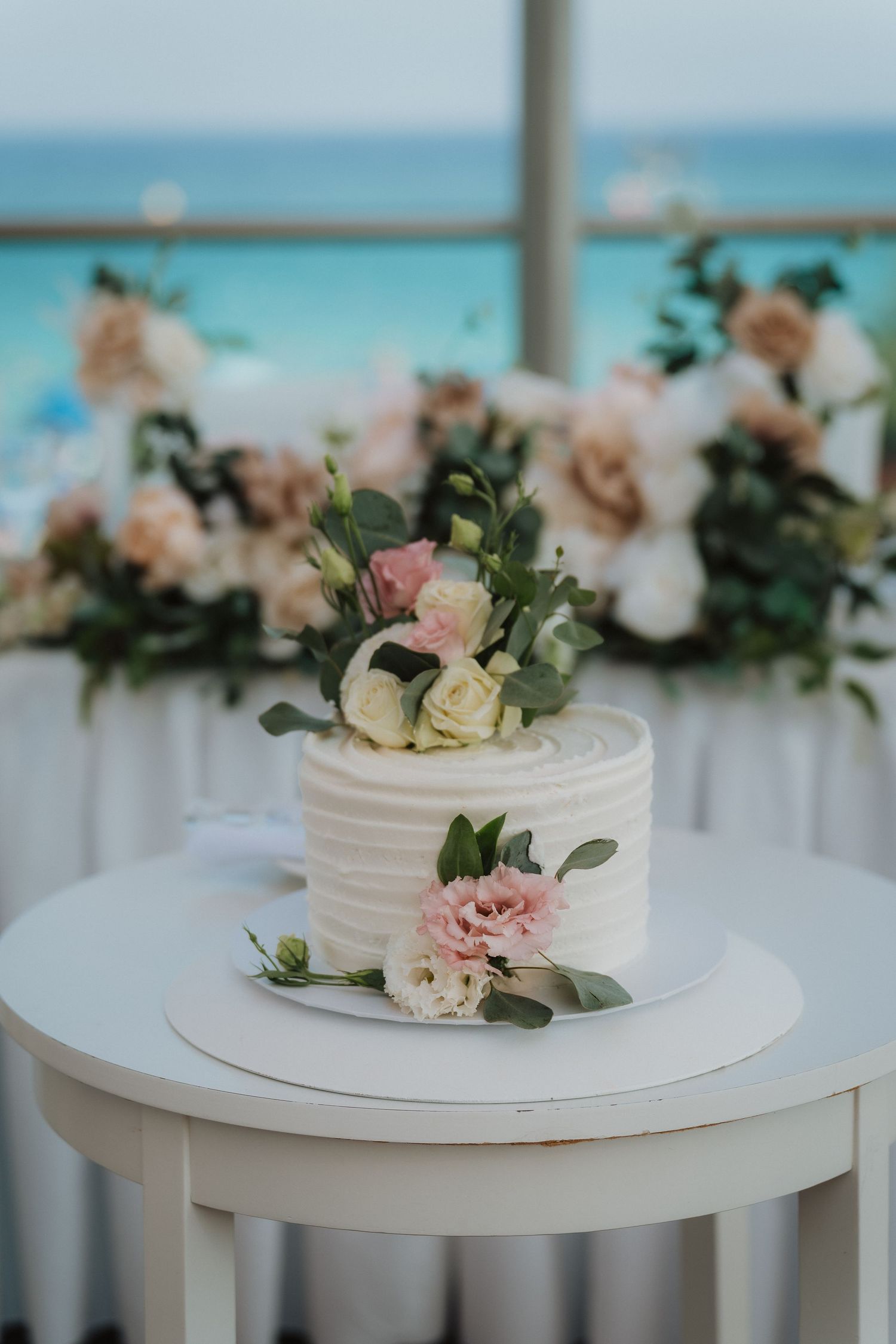 Elegant white wedding cake decorated with pink and cream roses on a round table with ocean views.