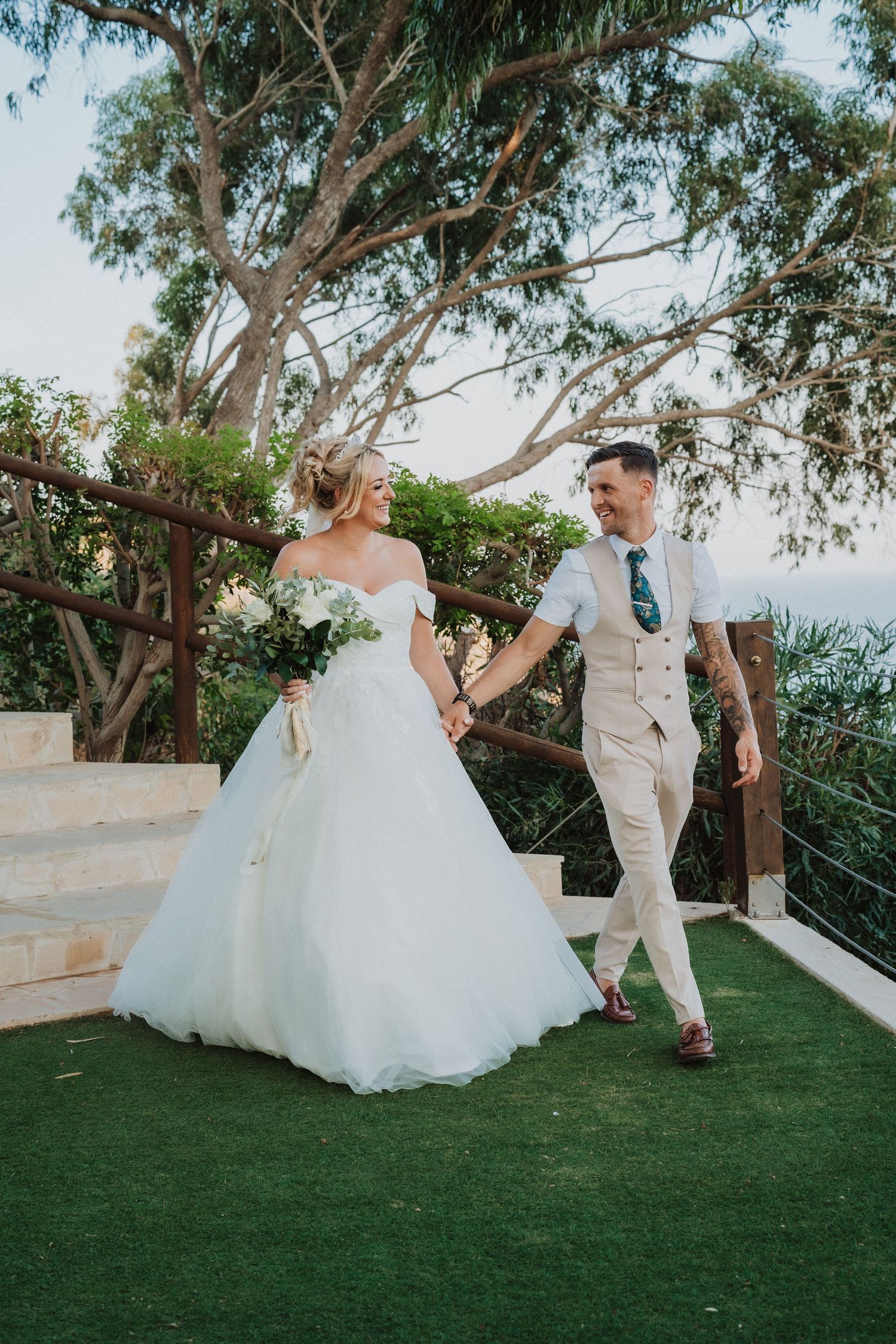 Newlyweds walk hand in hand on green lawn with eucalyptus trees in background.