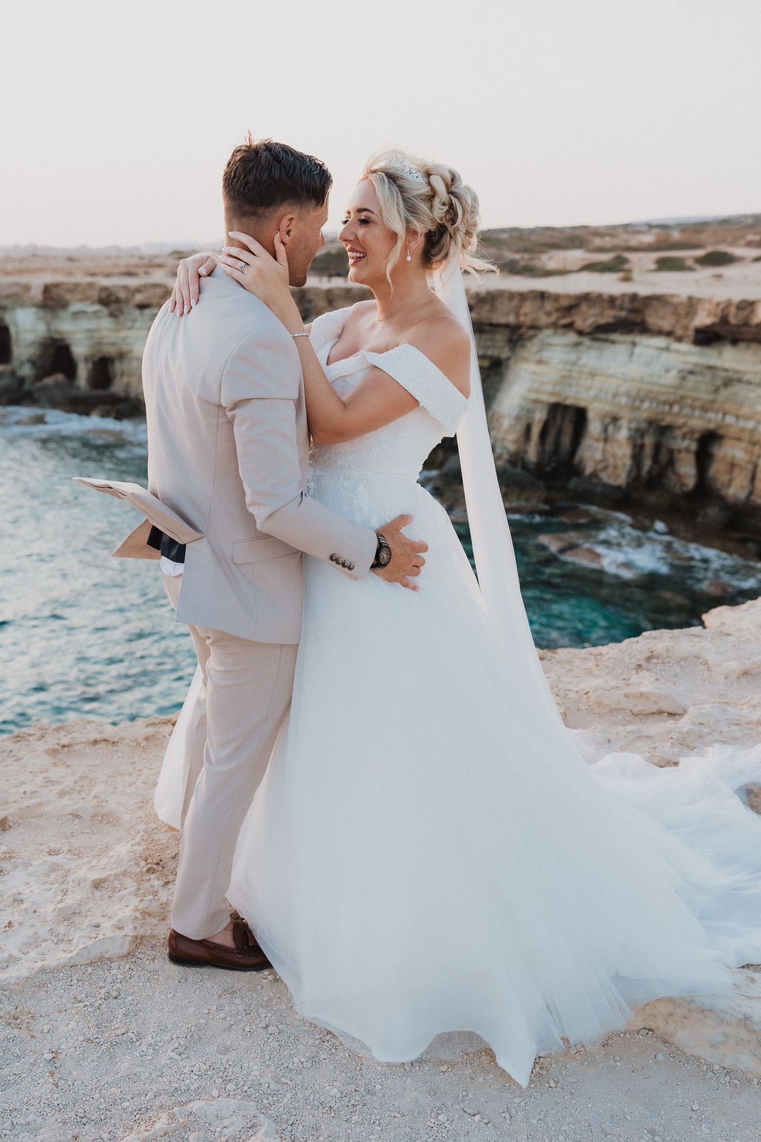 Newlyweds embrace on coastal cliffs overlooking the ocean in formal wedding attire.