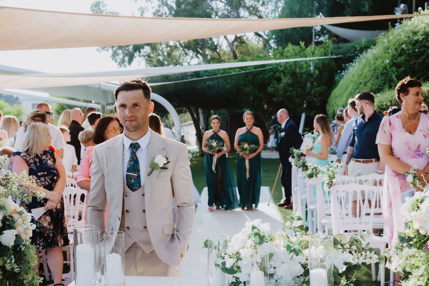 A wedding ceremony aisle decorated with white flowers and chairs under beige sail shades in a garden setting.