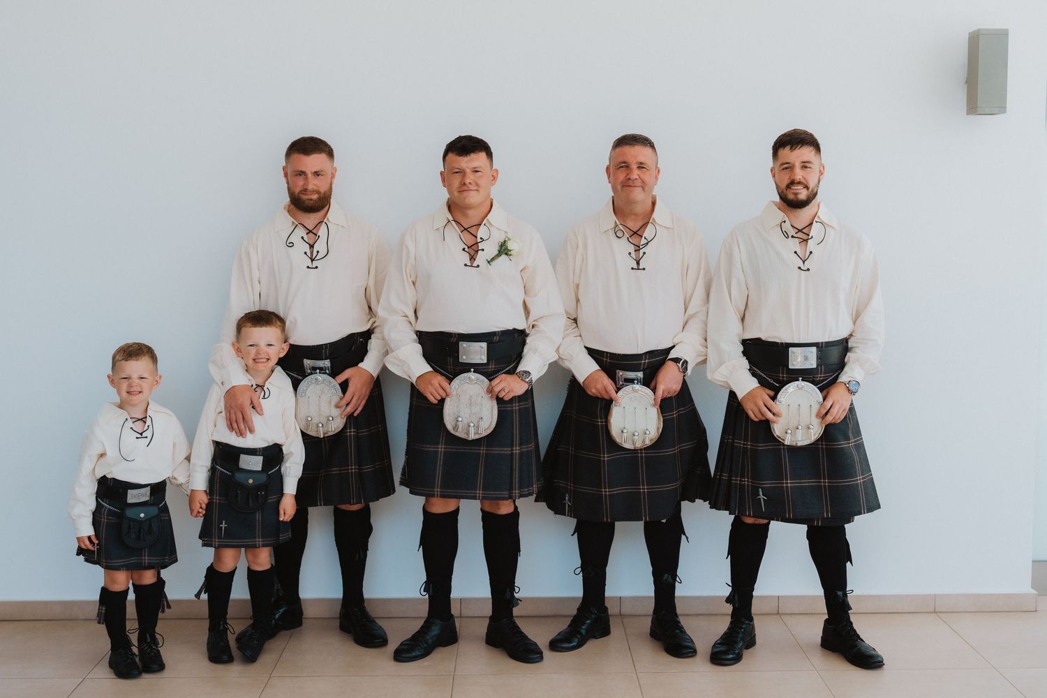 A group wearing matching traditional Scottish kilts and white shirts pose against a white wall.