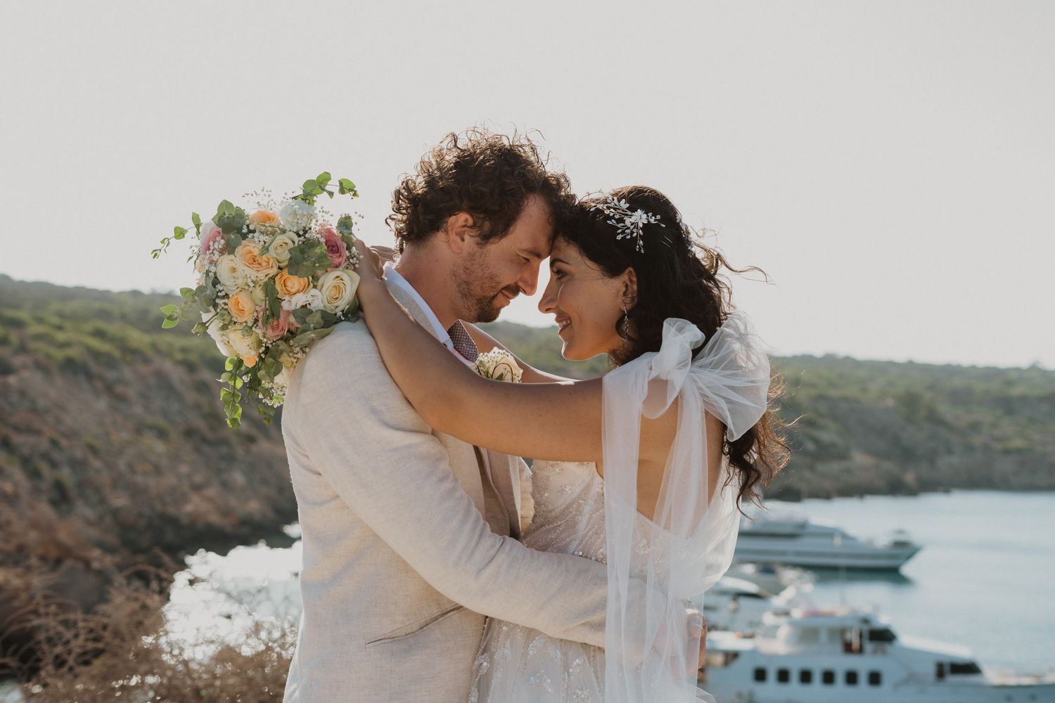 An intimate embrace between newlyweds on a Mediterranean cliffside with boats in the harbor below.