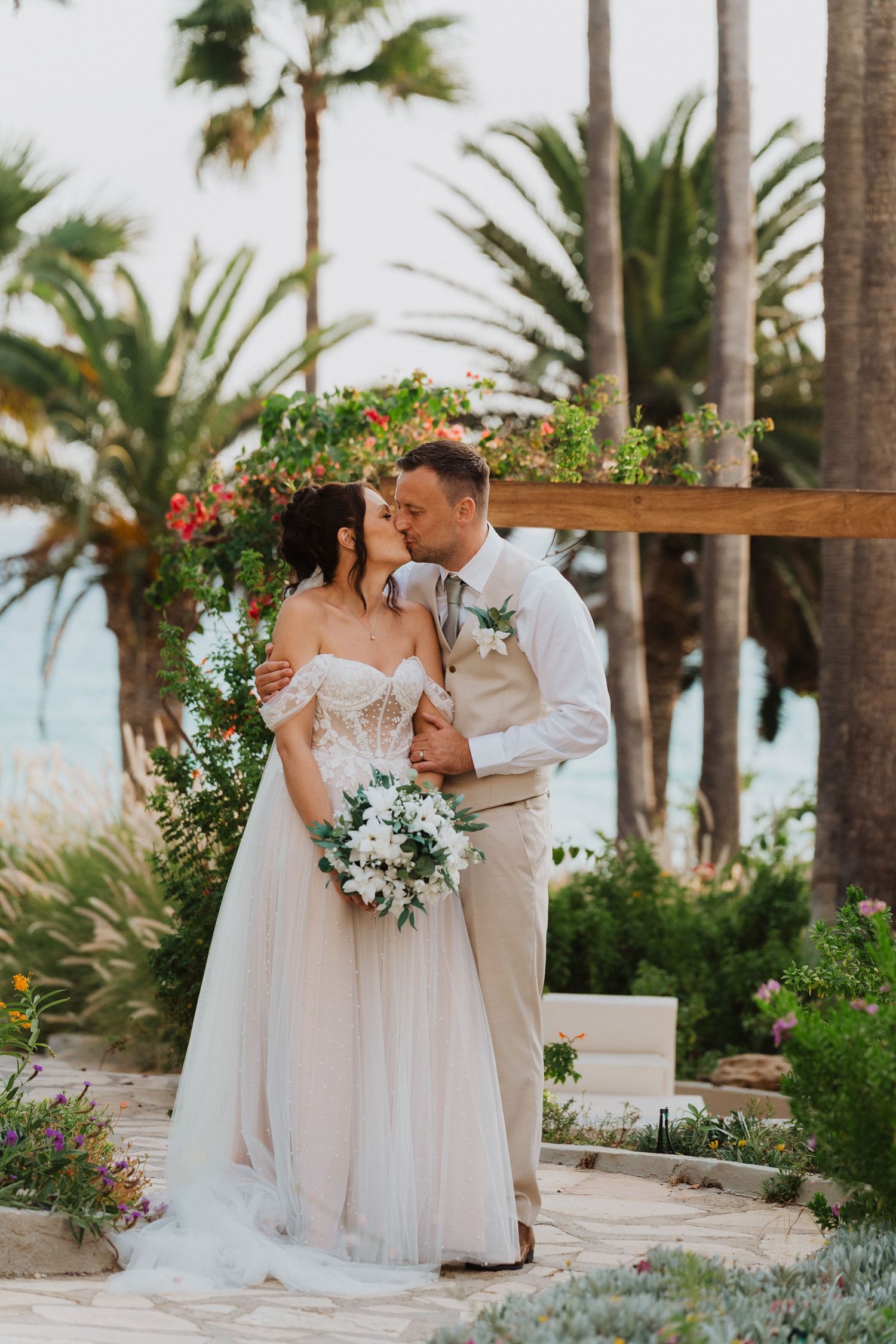 A romantic wedding couple embraces under a floral arch with palm trees and ocean views in the background.