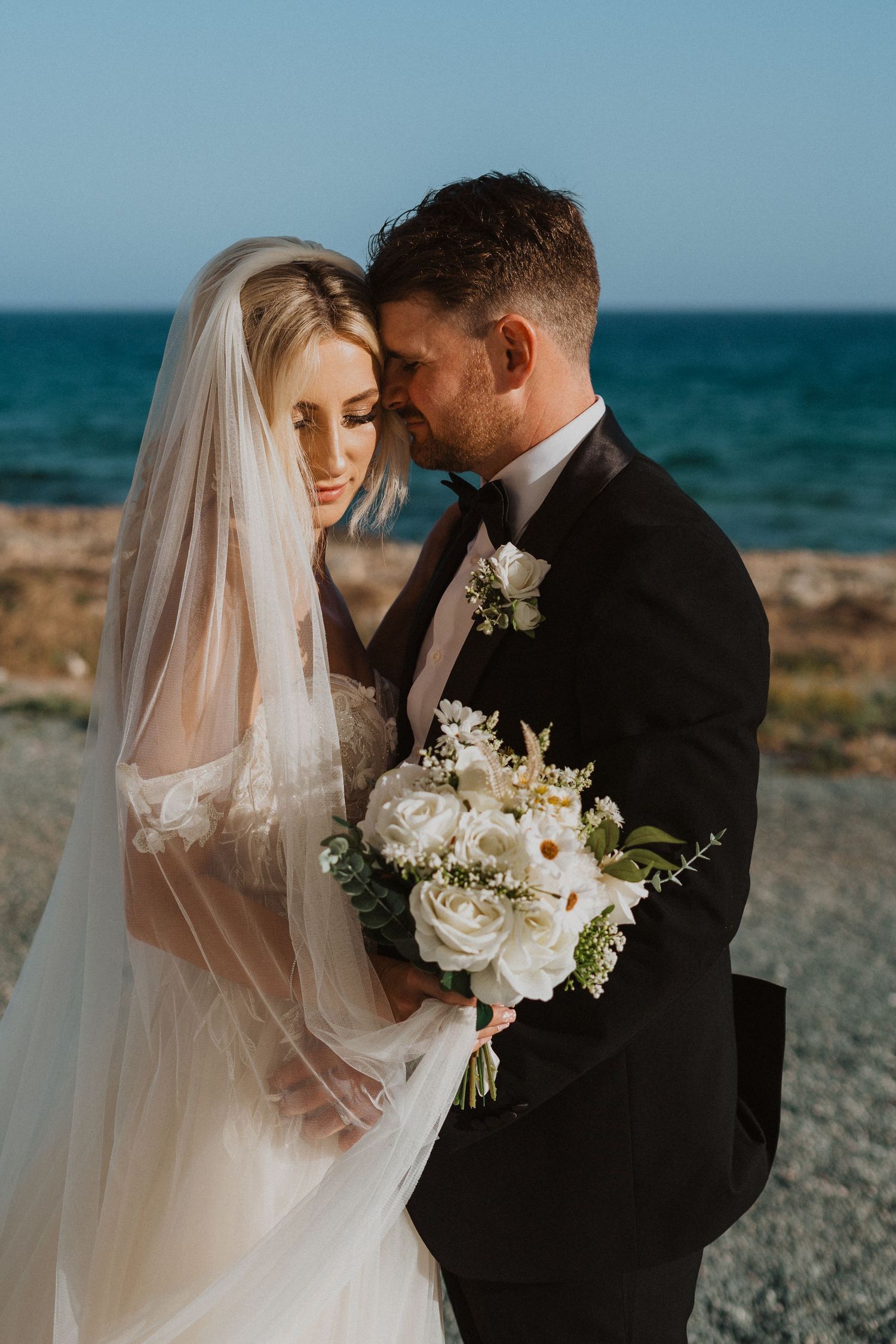 Intimate beachside wedding portrait featuring elegant white bouquet and flowing bridal veil.