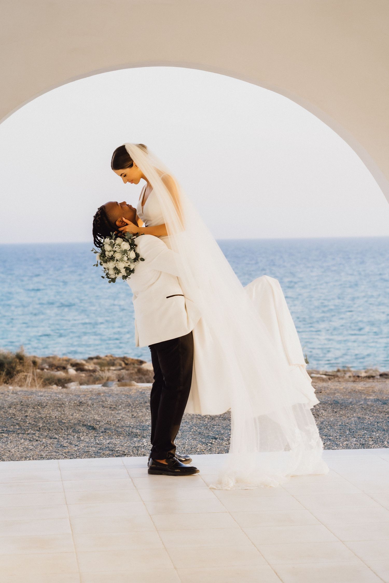 A romantic lift pose under a white Mediterranean arch with ocean views and flowing veil.
