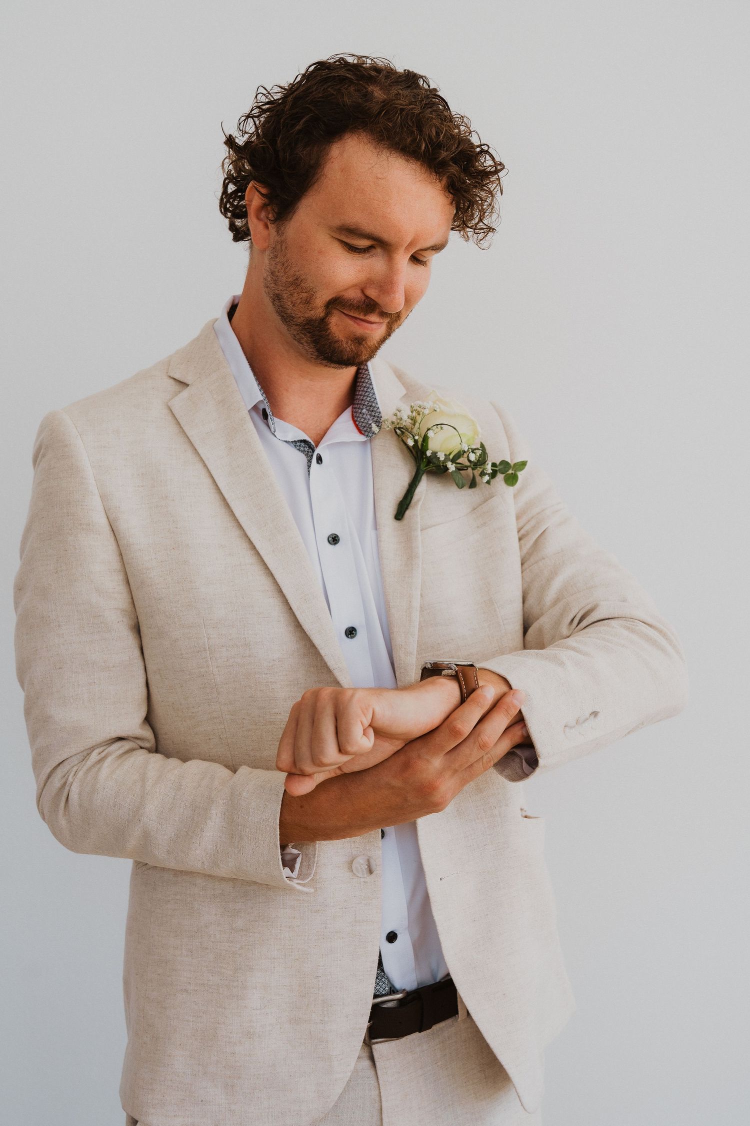 Groom in beige suit checks time on watch while wearing boutonniere.