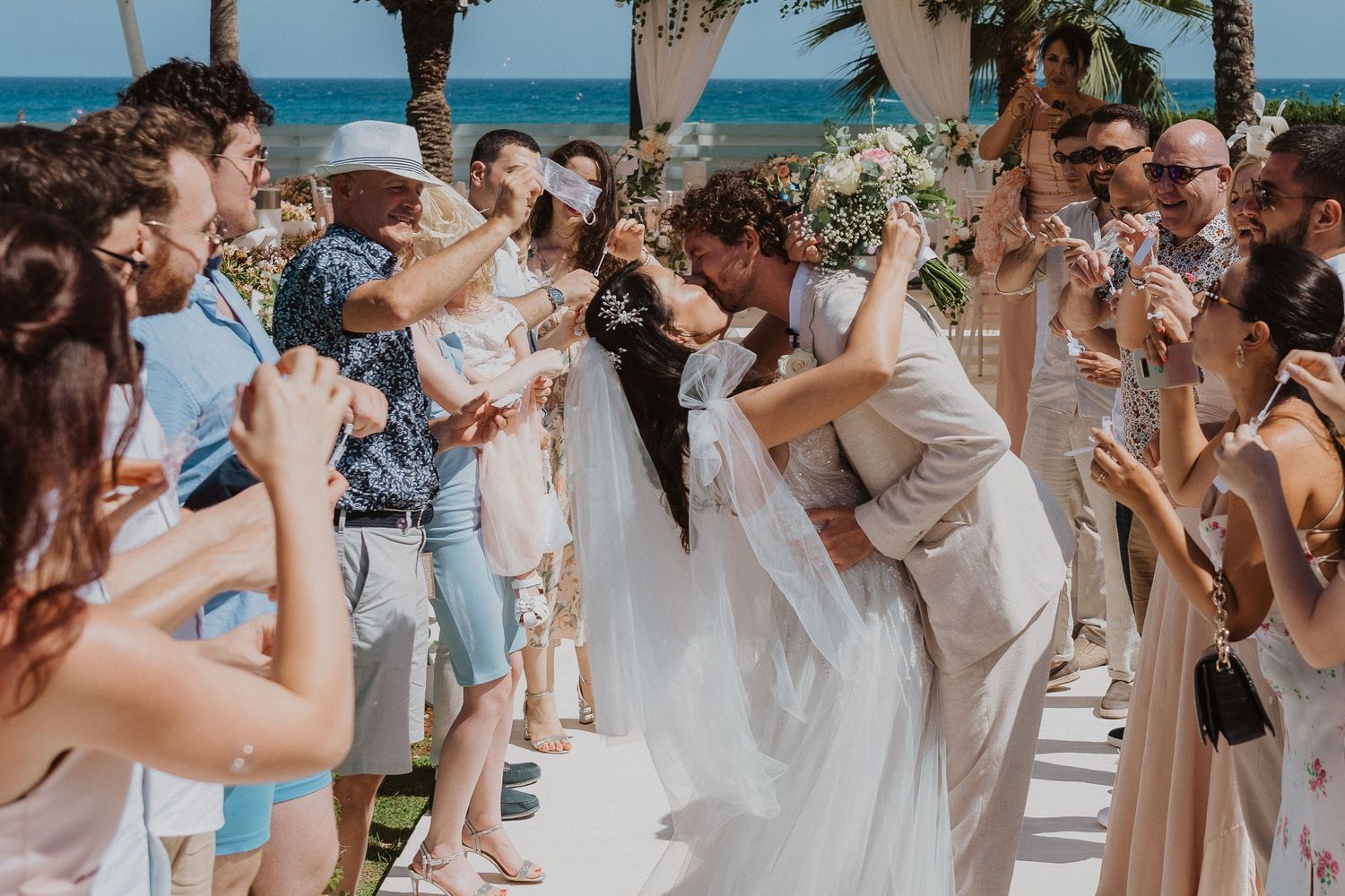 Wedding party walks down aisle under beige canopy by ocean as guests throw confetti.