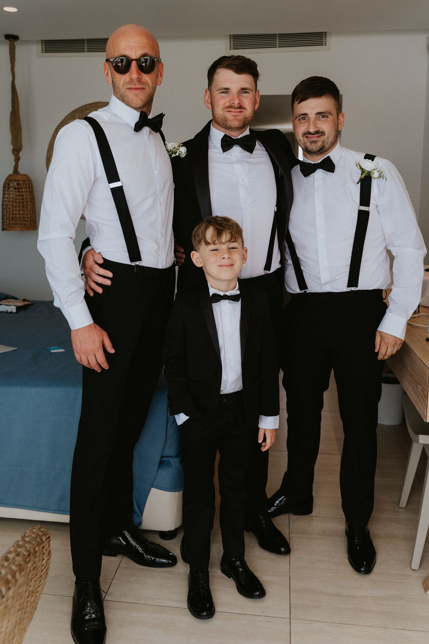 A formal wedding party photo featuring men in black and white attire with suspenders and bowties.