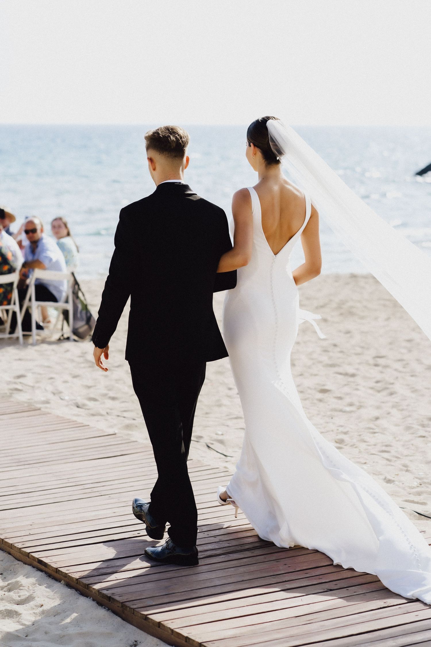 A couple walks down a wooden boardwalk on a beach in wedding attire.