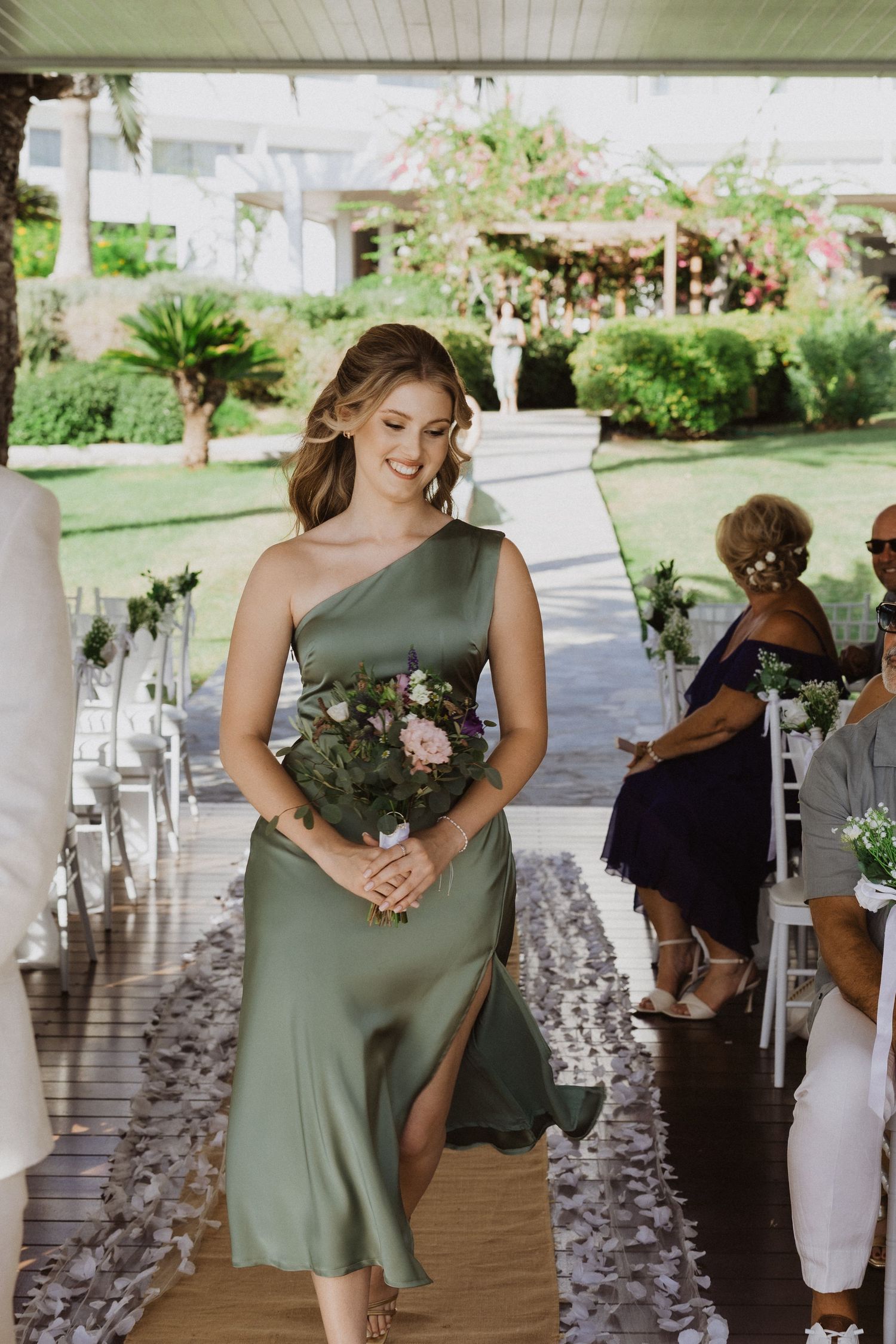Bridesmaid walks down aisle decorated with purple petals on wooden deck.