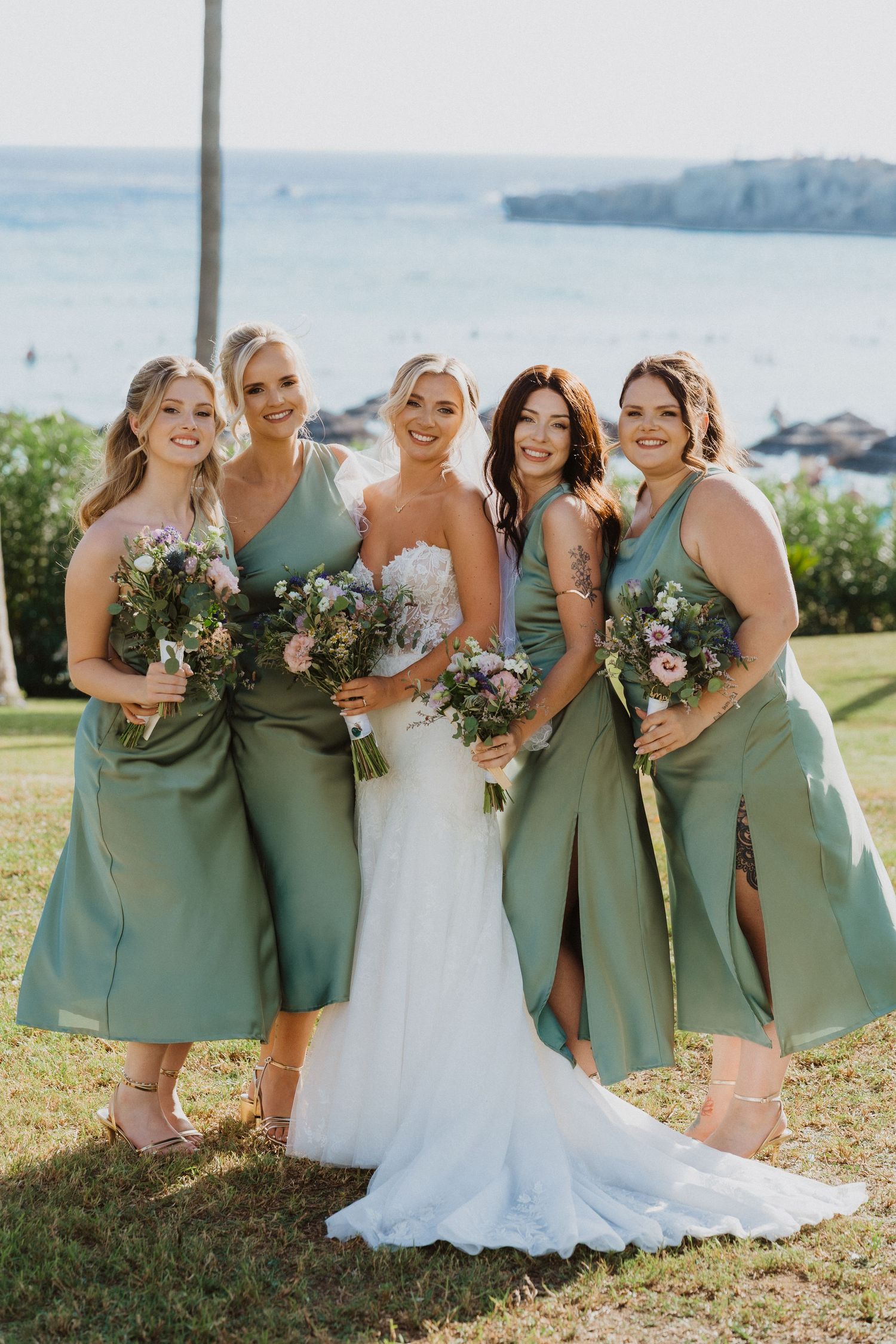 Group of bridesmaids in sage green dresses stand together on coastal lawn with ocean view.