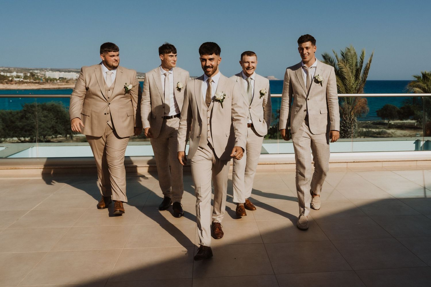 Five groomsmen in tan suits walking along seaside venue.